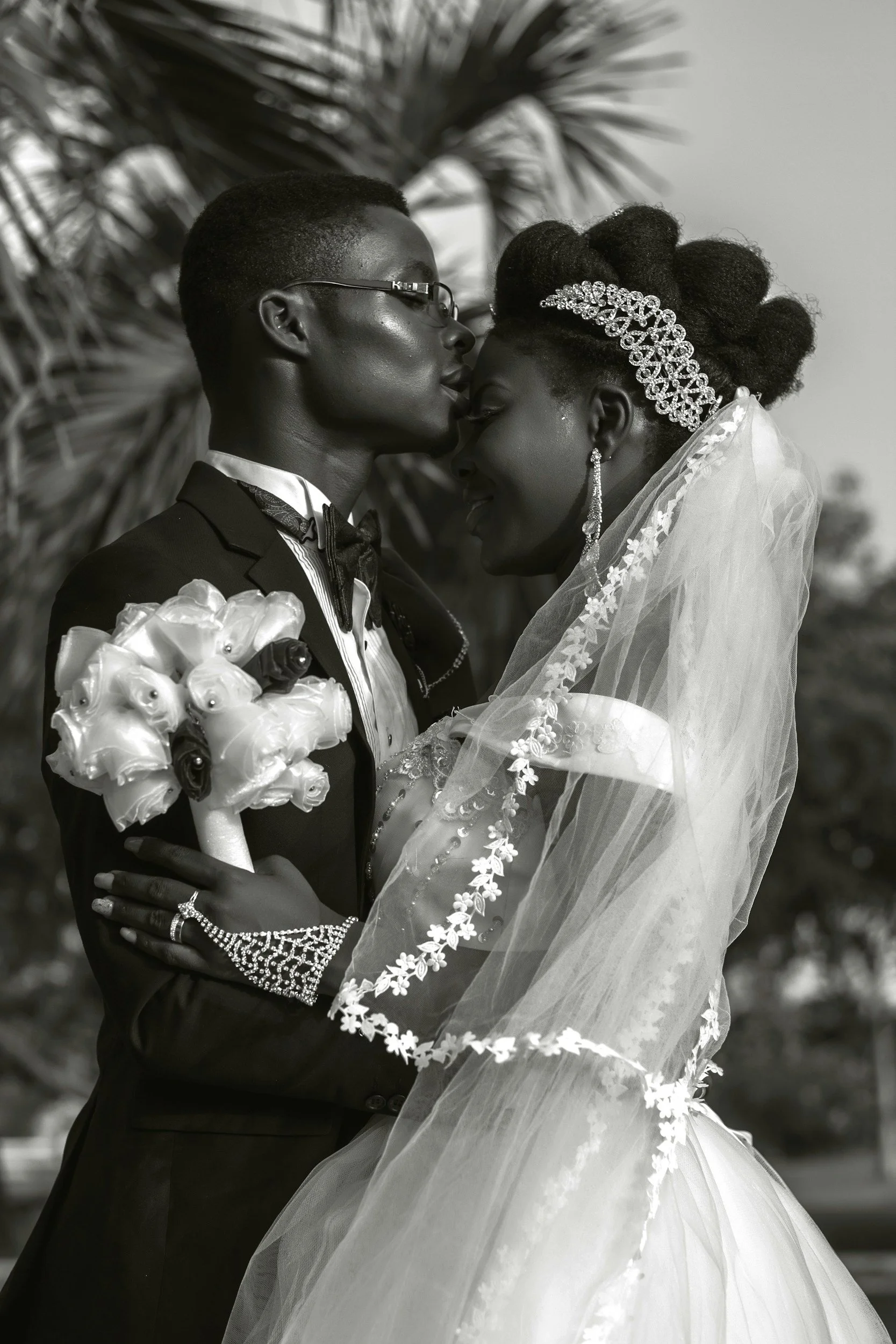 A black and white photo of a couple in wedding attire, embracing each other closely with their foreheads nearly touching, outdoors with blurred trees in the background.