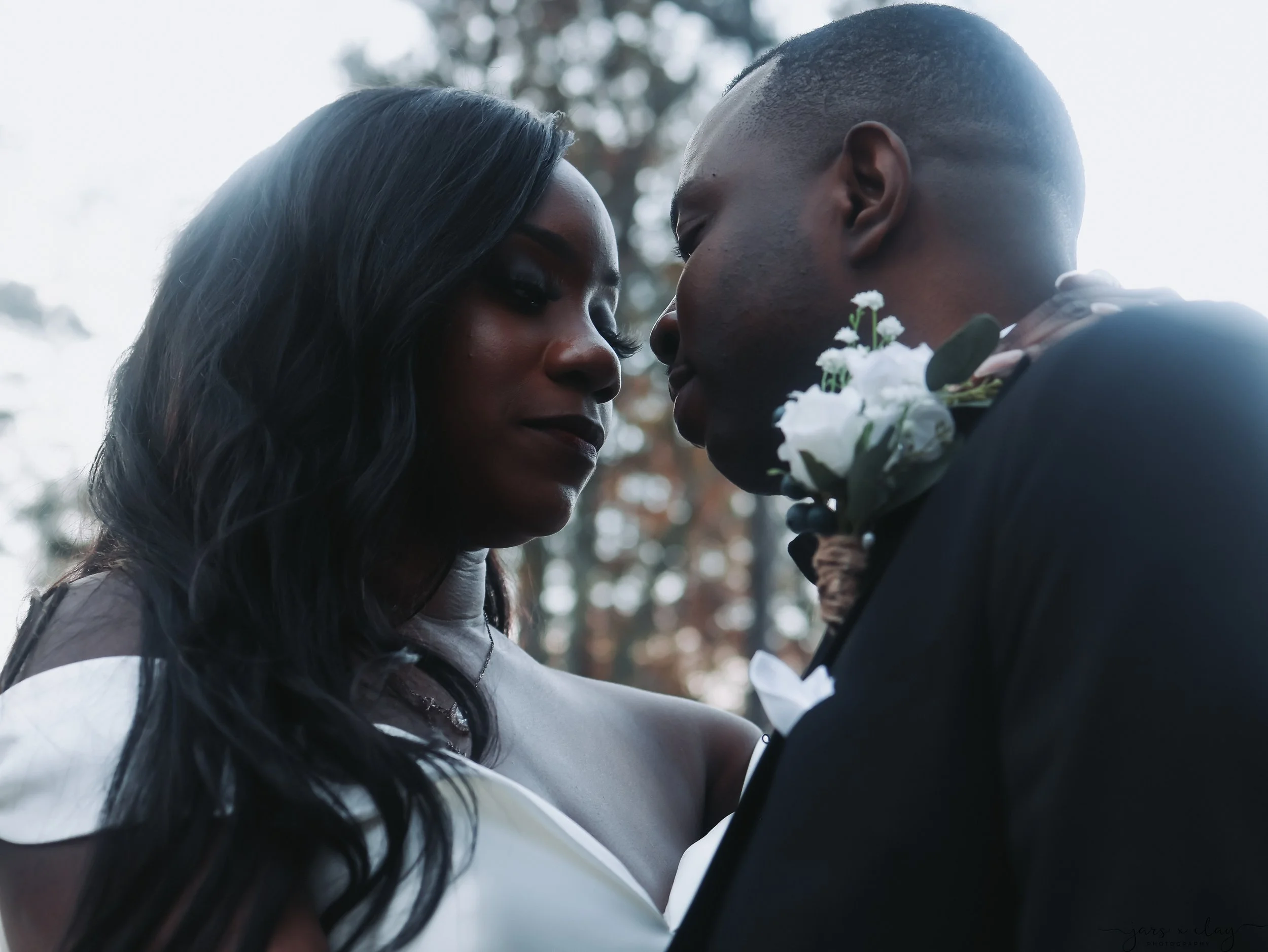 A couple facing each other closely outdoors, with the woman in a white dress and the man in a suit, holding white flowers.
