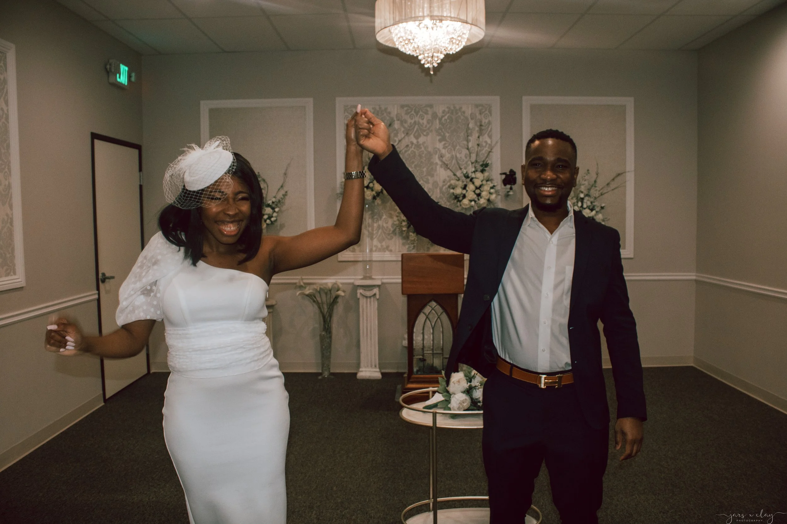 A couple is dancing at a celebration in a decorated room with floral arrangements and a chandelier. The woman is wearing a white dress with a peplum detail and a fascinator, while the man is dressed in a black jacket and white shirt. They are smiling and holding hands up in a dance pose.