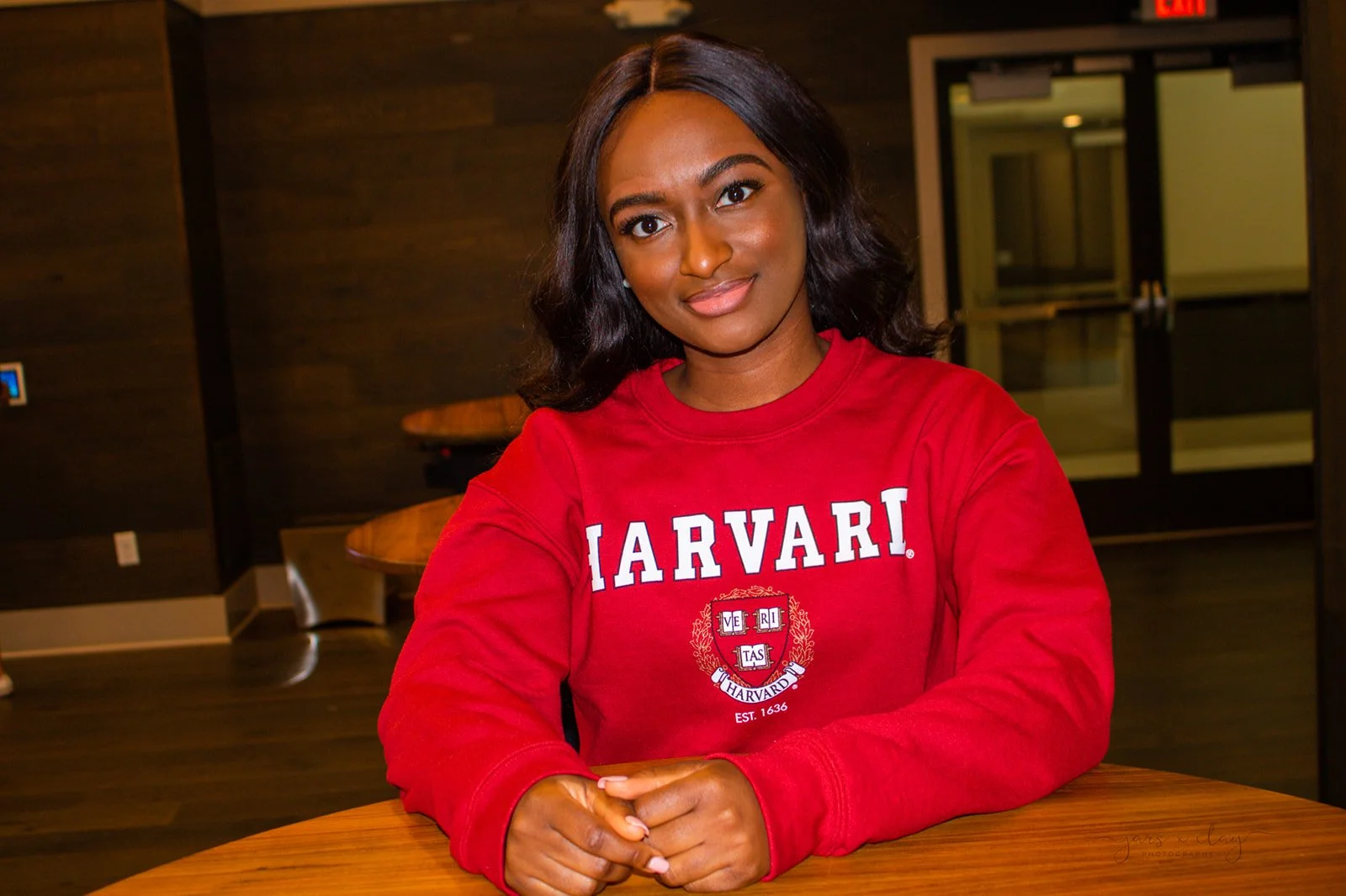 A young woman with dark hair and brown skin wearing a red Harvard sweatshirt, sitting at a wooden table in an indoor setting with dark walls and glass doors in the background.