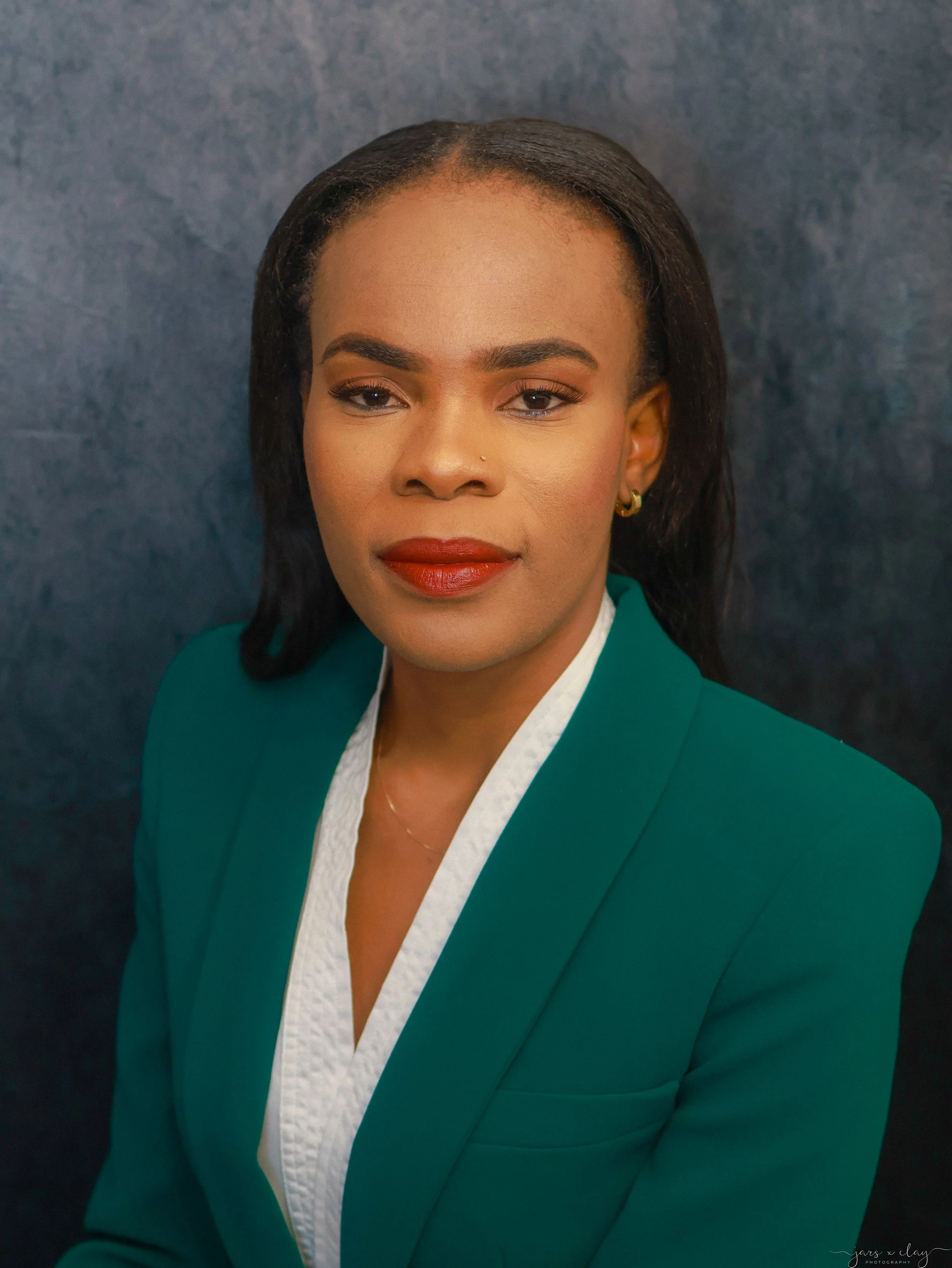 A portrait of a woman with dark hair, wearing a green blazer and white top, set against a dark textured background.