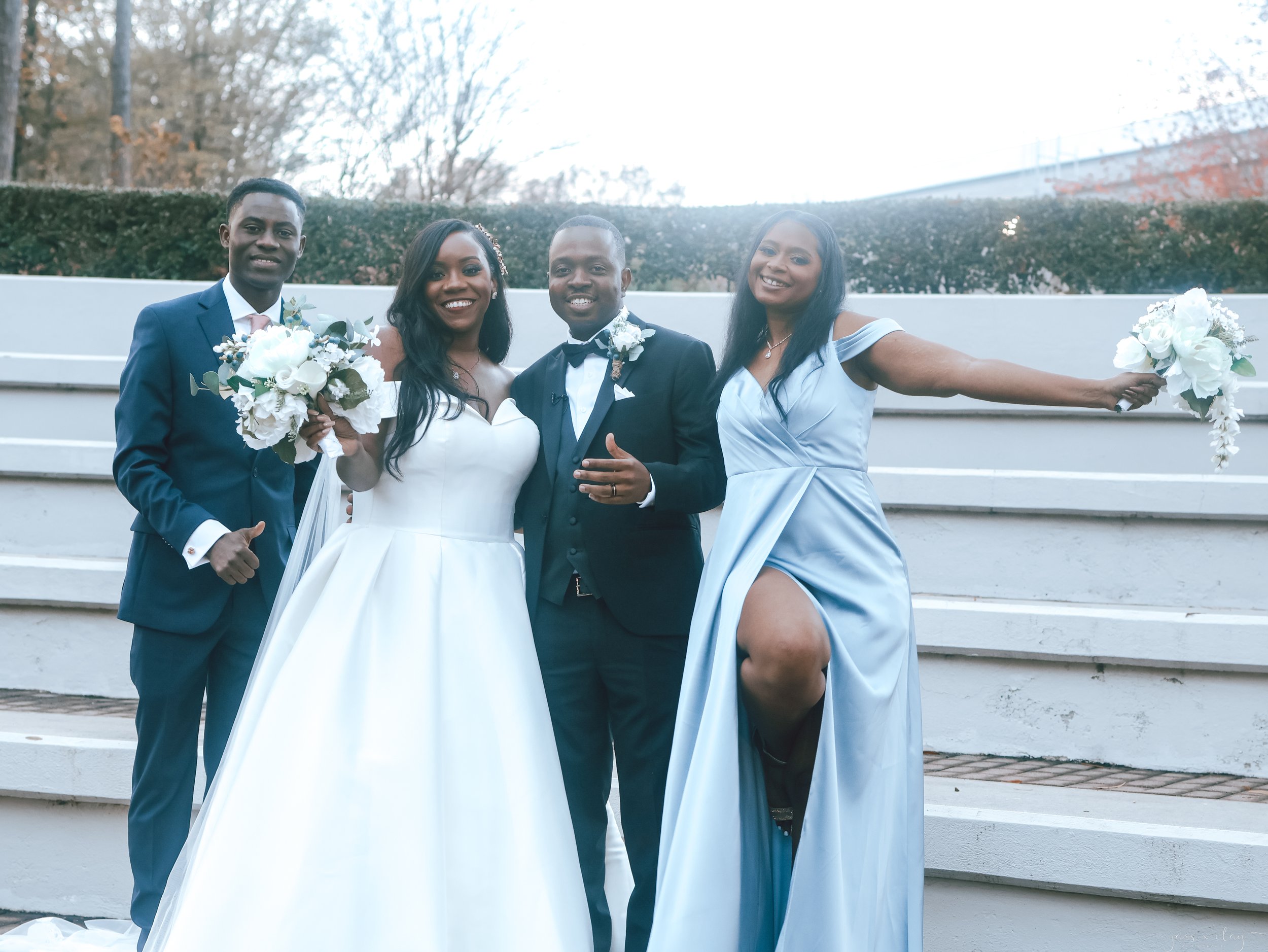 A group of four people, dressed in formal attire, smiling and posing together outdoors, with some holding bouquets of white flowers. The background features trees and a modern, white concrete structure.
