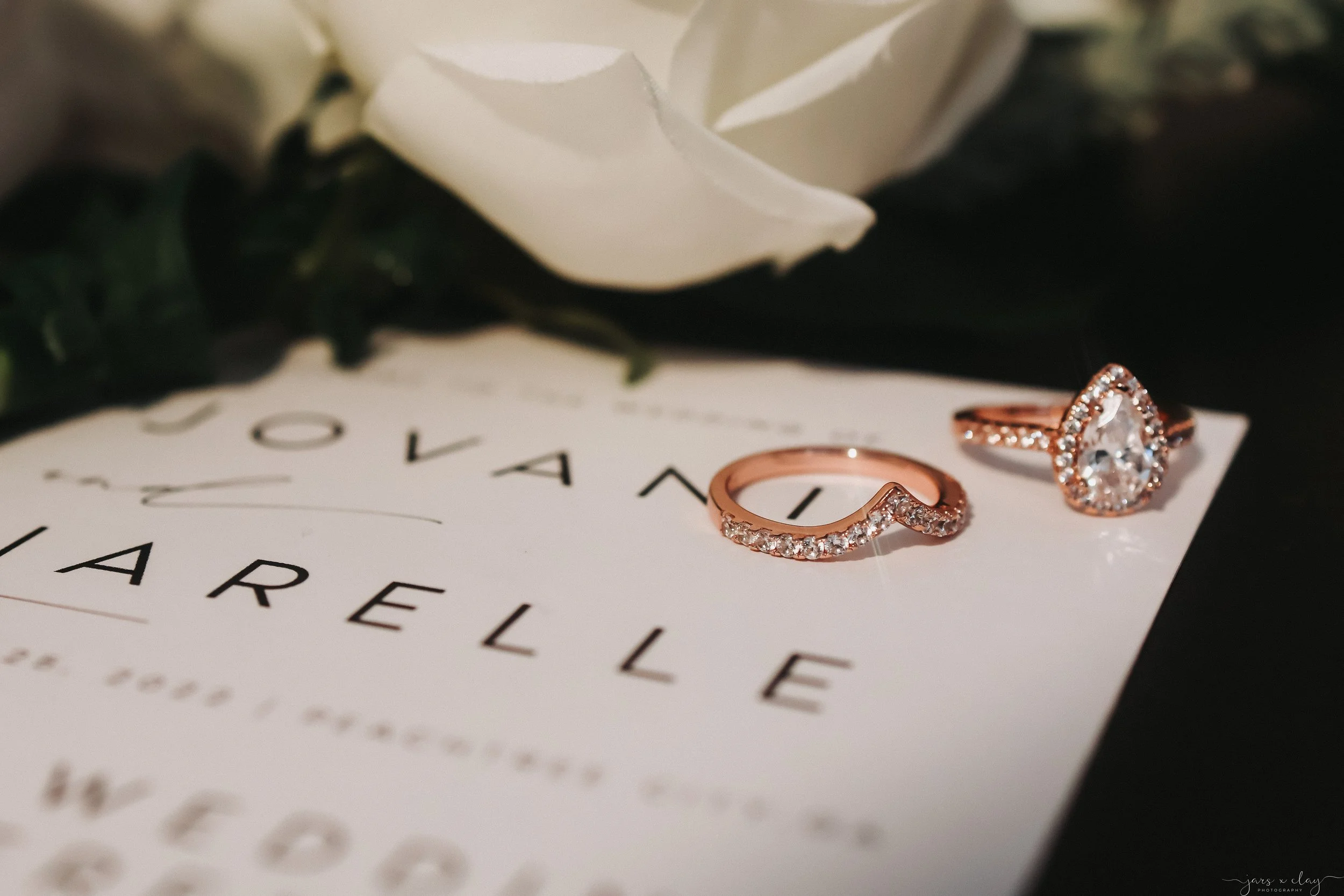 Close-up of a white rose, two rose gold rings with gemstones, and part of an invitation with the text 'WEDDING REELLE'.