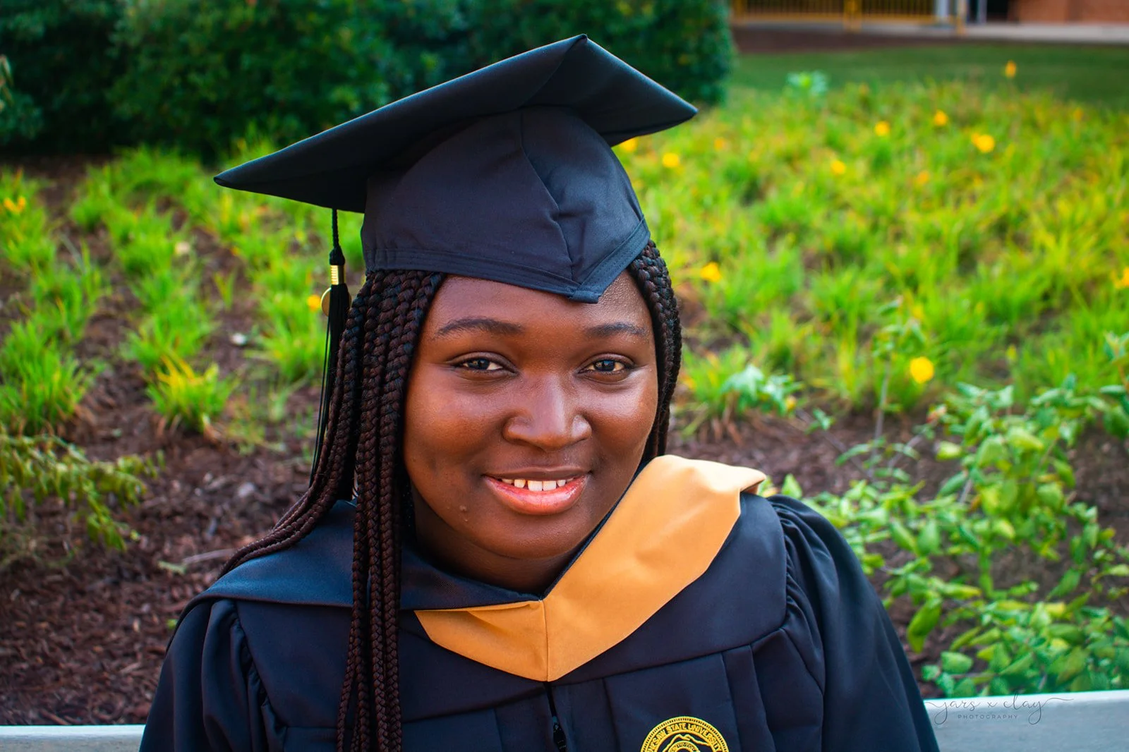 Young woman wearing a graduation cap and gown, smiling outdoors near green bushes and yellow flowers.
