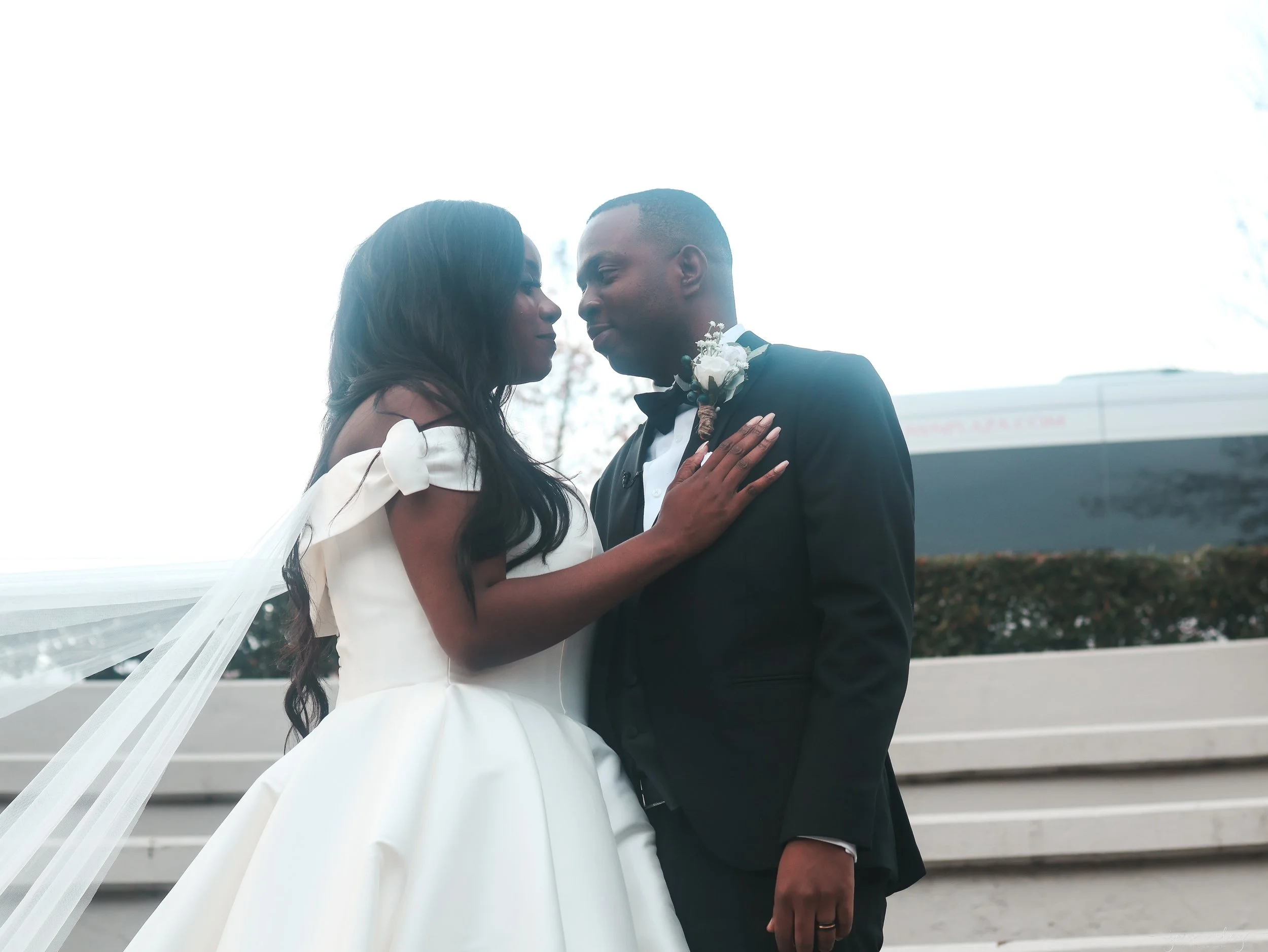 A bride and groom sharing an intimate moment, facing each other outdoors, with the bride in a white gown and a veil, and the groom in a black tuxedo with a boutonniere.