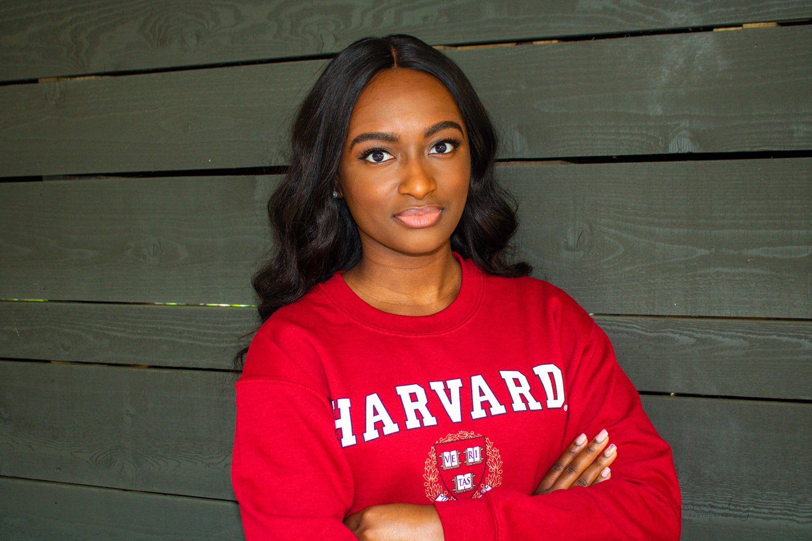 A young African American woman with long dark hair, wearing a red Harvard sweatshirt, standing with arms crossed in front of a dark wooden wall.