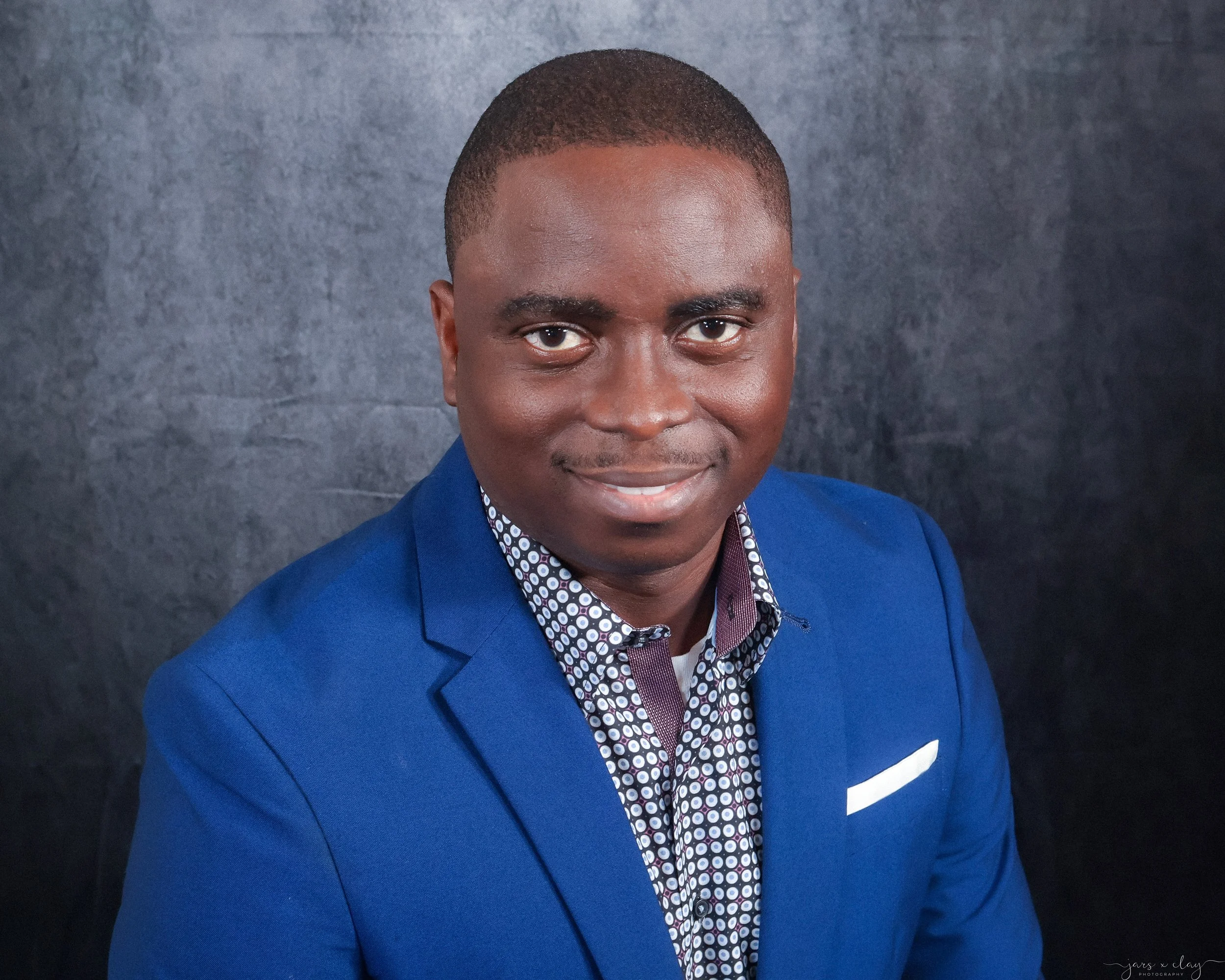 Portrait of a man wearing a bright blue blazer and a patterned shirt, smiling, against a dark textured background.