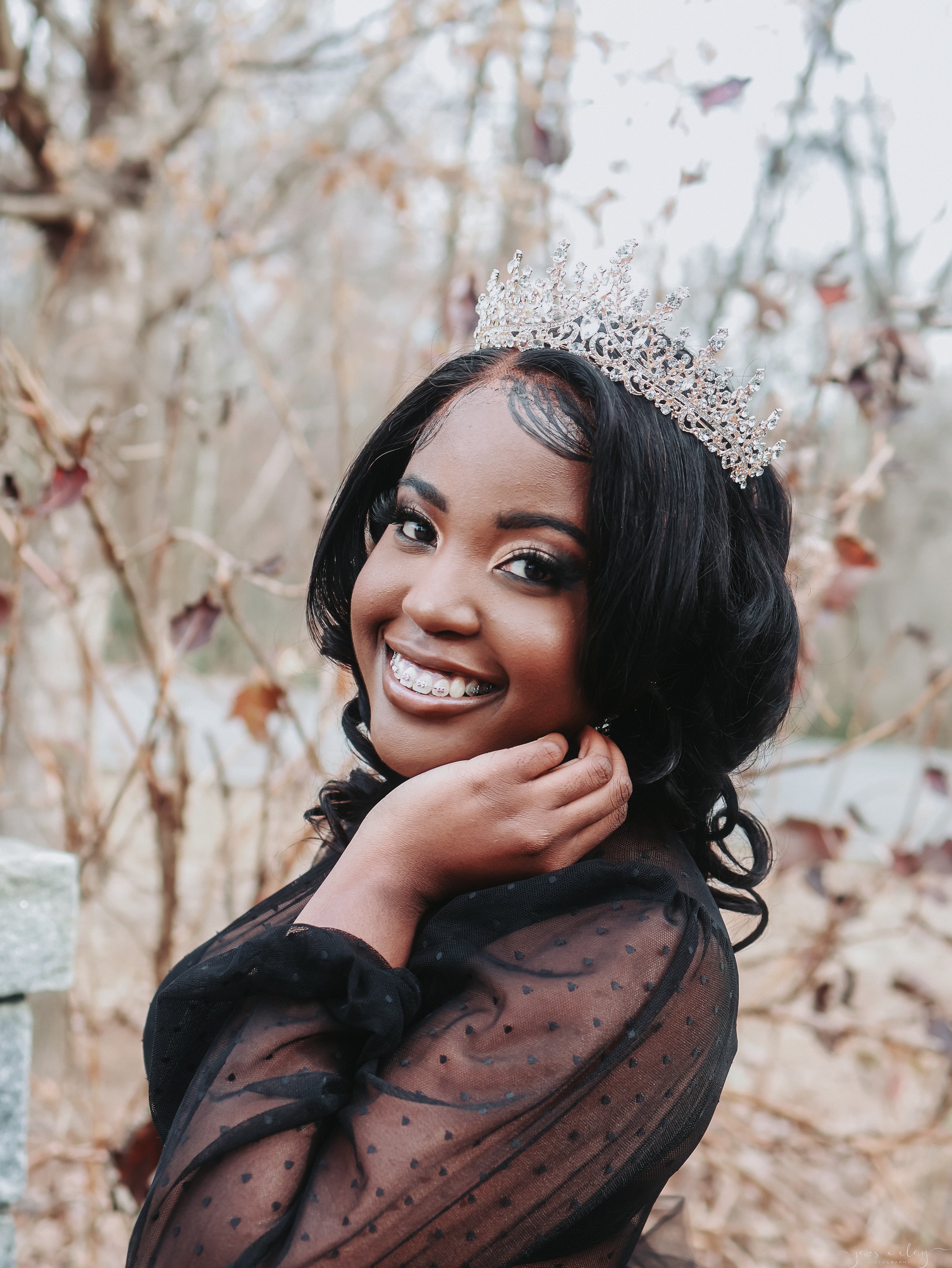 A woman with dark hair wearing a decorative tiara smiles outdoors in front of bare trees.