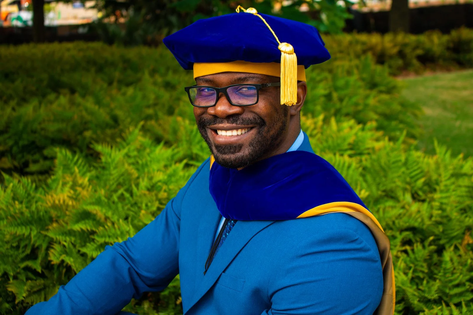 Graduate wearing a blue cap and gown with a yellow tassel, smiling outdoors with greenery in the background.