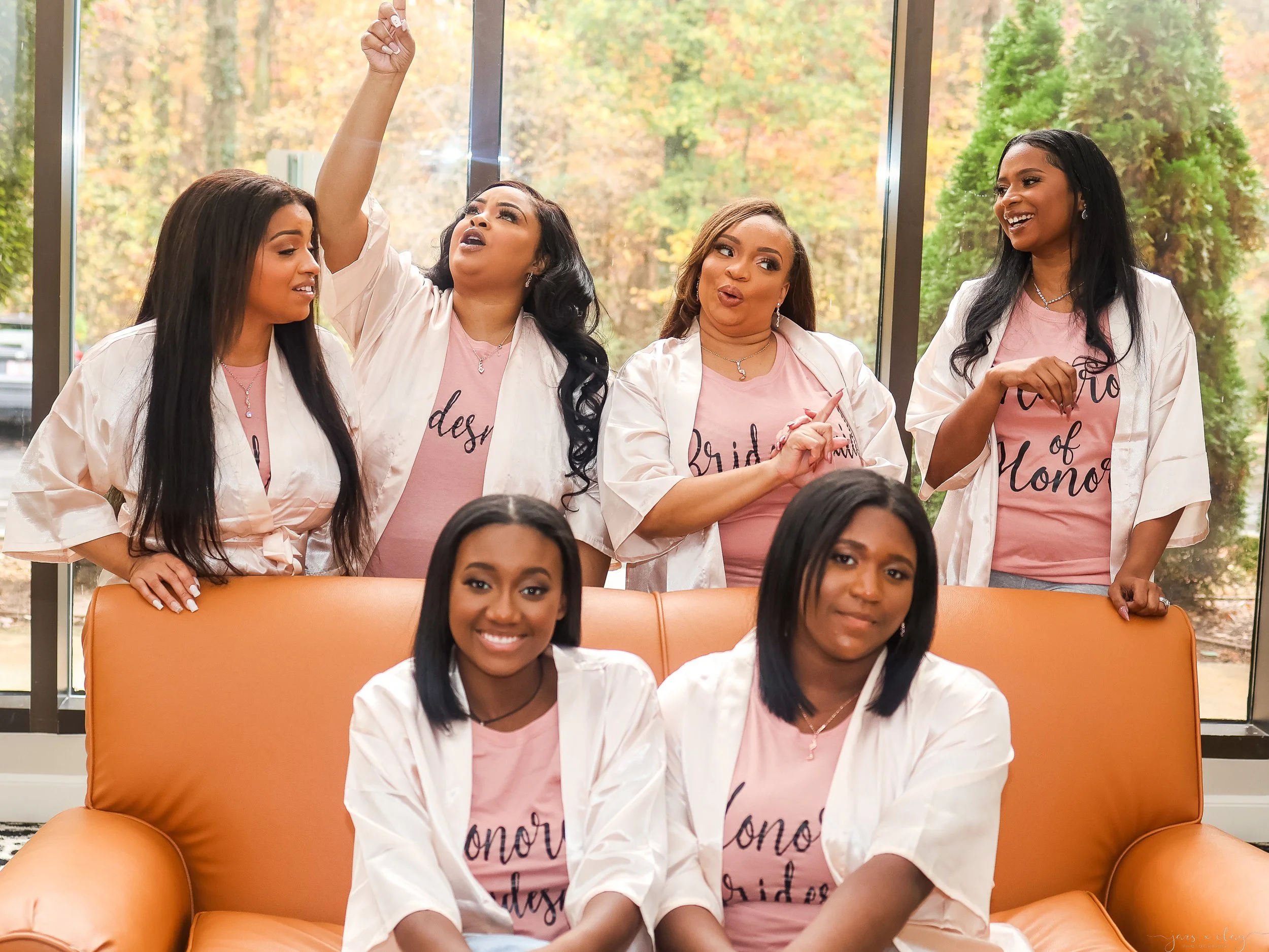 Group of four women wearing pink shirts and white robes, celebrating together indoors, with large windows and a fall landscape in the background.