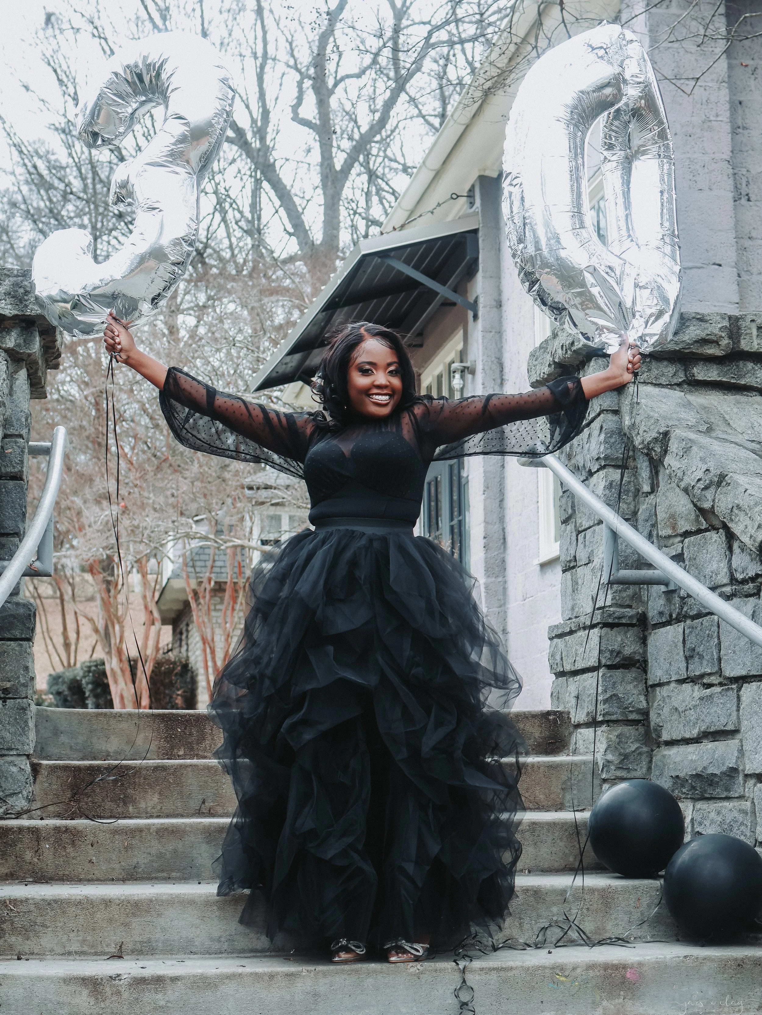 Woman in a black, tulle ball gown holding two silver balloons shaped as the numbers 2 and 0 outside on stone steps with a stone wall and leafless trees in the background.