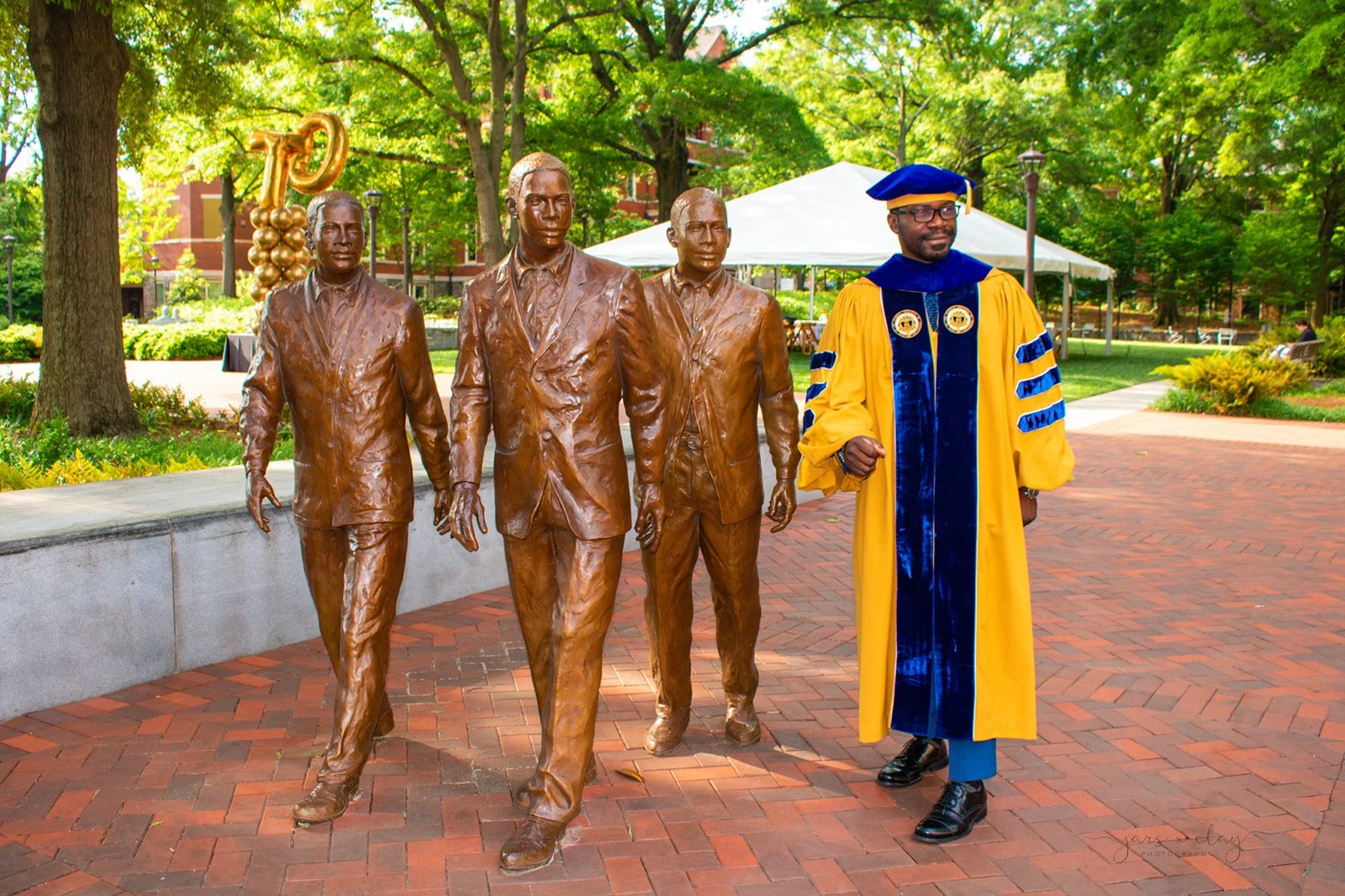 A person in academic regalia walking past a bronze monument of four men in suits. The background shows green trees, a white tent, and brick pathways.