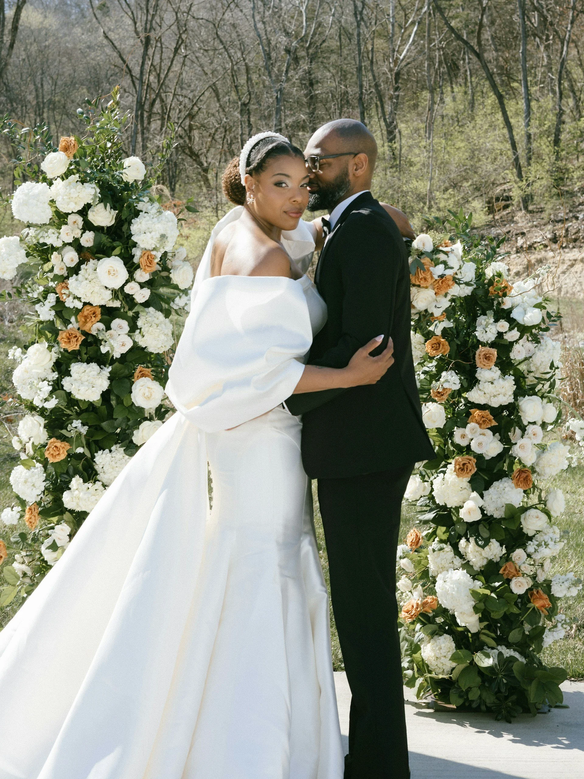 A newlywed couple in wedding attire stands closely together outdoors, with the bride in a white wedding gown and the groom in a black tuxedo. They are in front of floral arrangements with white and peach flowers and surrounded by trees.
