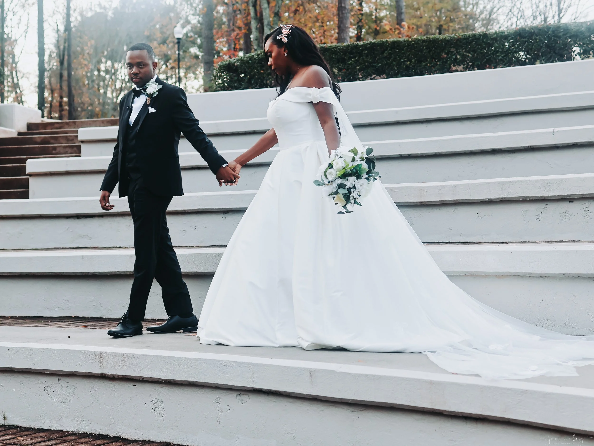 A bride and groom holding hands on outdoor steps. The bride is wearing a white off-the-shoulder wedding dress and holding a bouquet of white flowers with greenery. The groom is dressed in a black tuxedo with a white shirt and a black bow tie. They ar