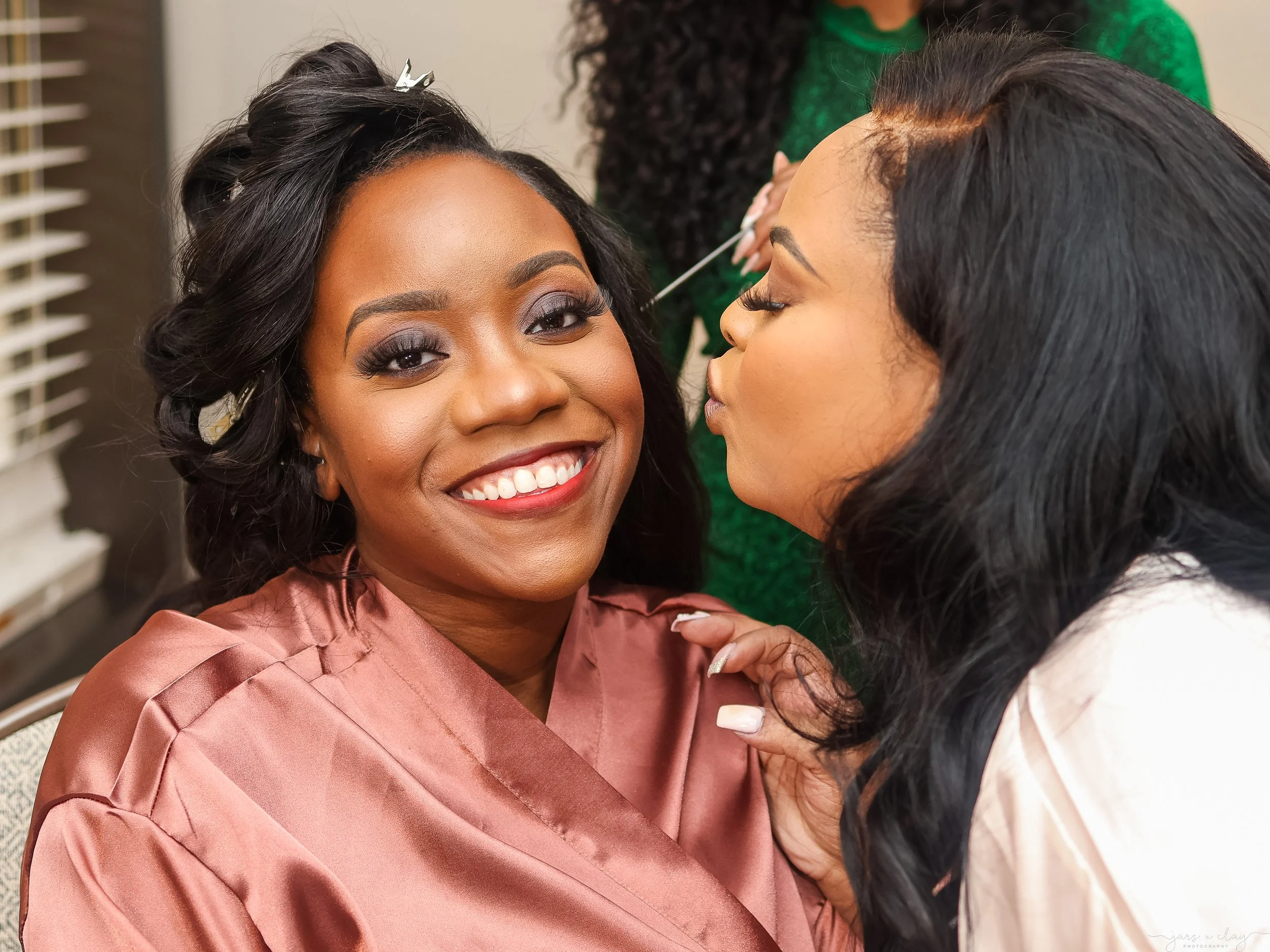 A woman with dark hair styled in waves, smiling with white teeth, wearing a shiny satin pink dress with puffed sleeves, and holding her hand near her chest. Another woman with black hair and curly hair extending to her shoulders is applying makeup wh