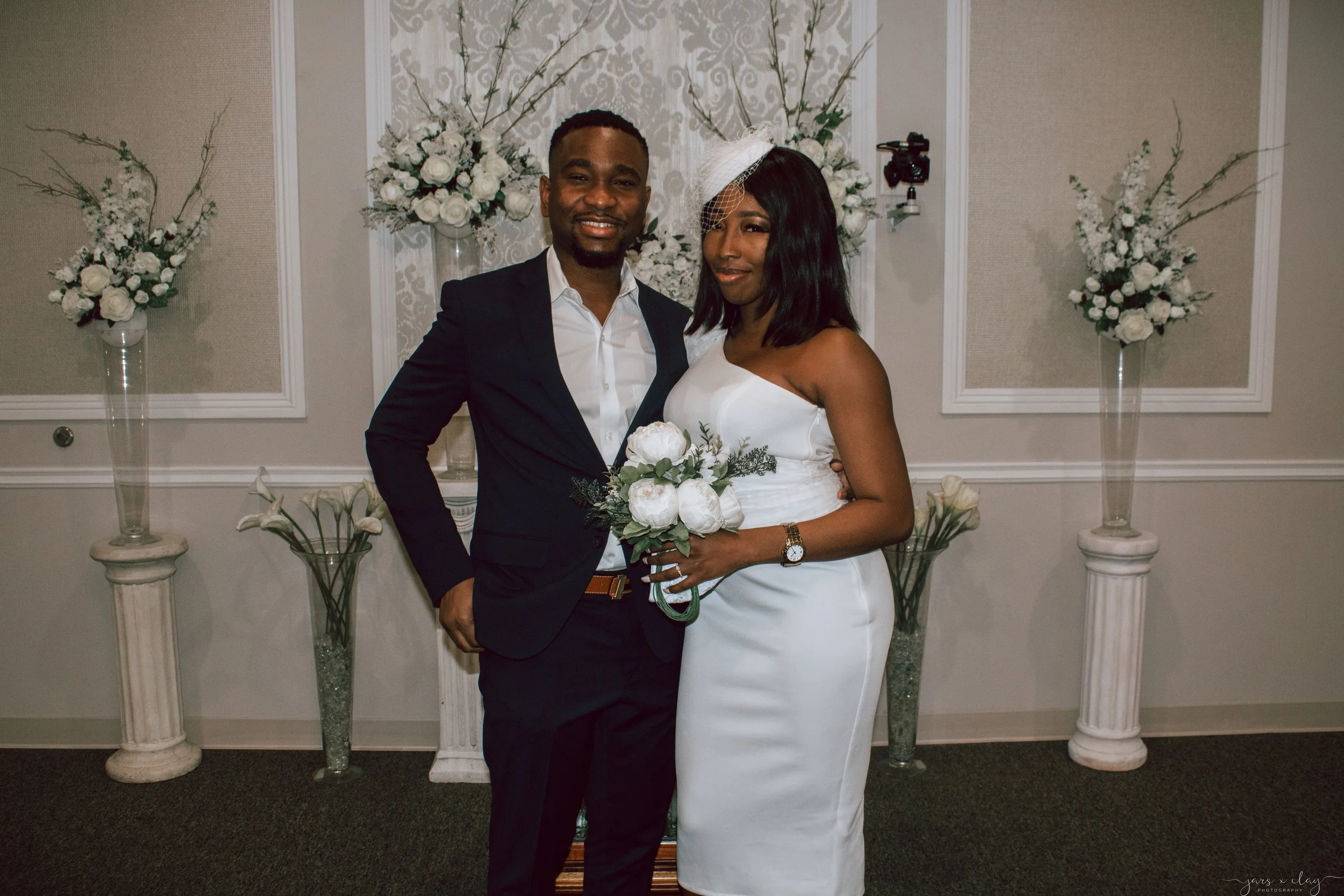 A bride and groom standing side by side smiling at the camera in front of a floral backdrop with white roses and greenery, in a wedding setting.