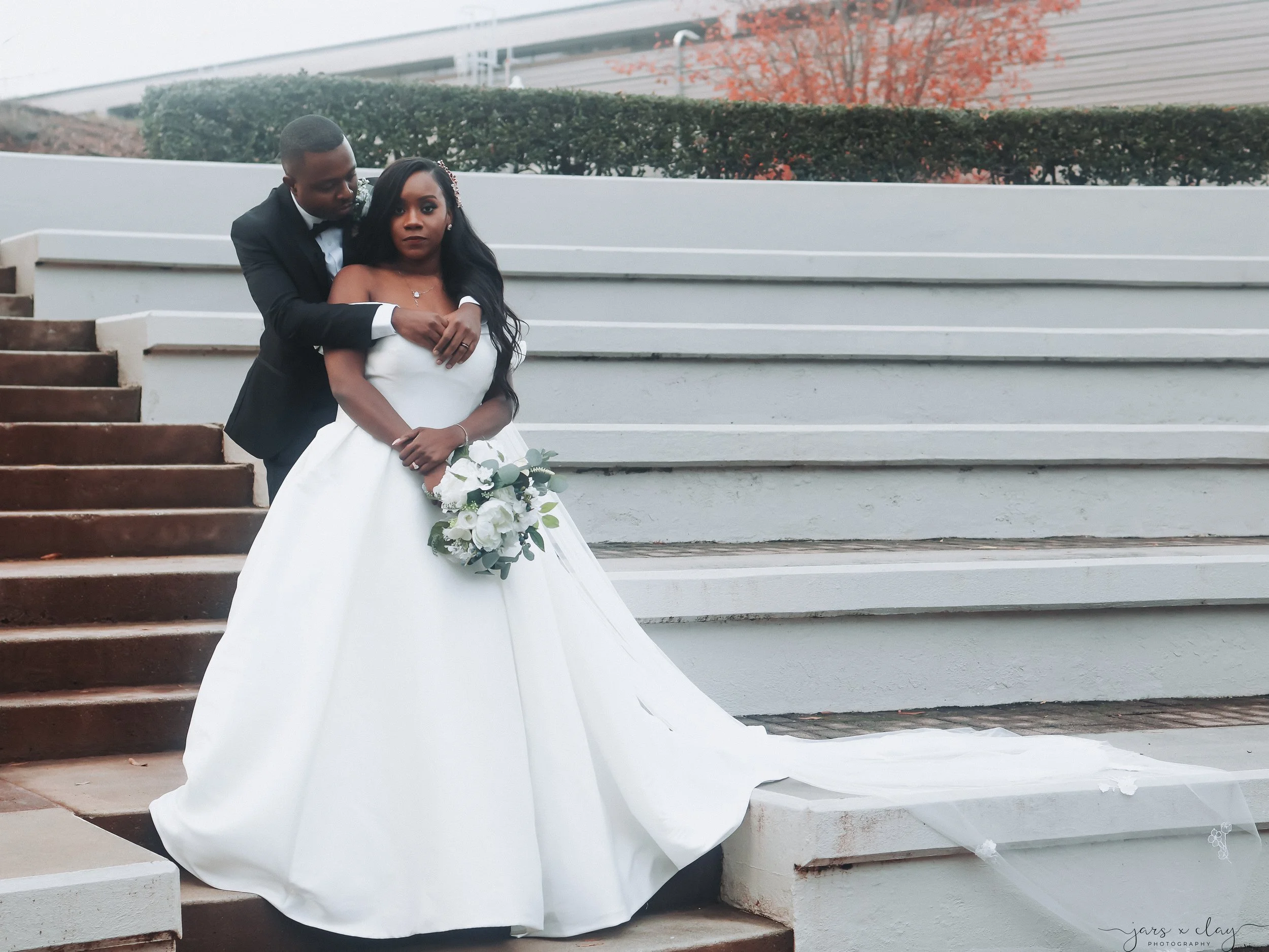 A bride and groom standing on stairs outside. The groom embraces the bride from behind as they pose for a photo. The bride is wearing a white wedding gown and holding a bouquet of white flowers. The background features a modern building, a tree with 