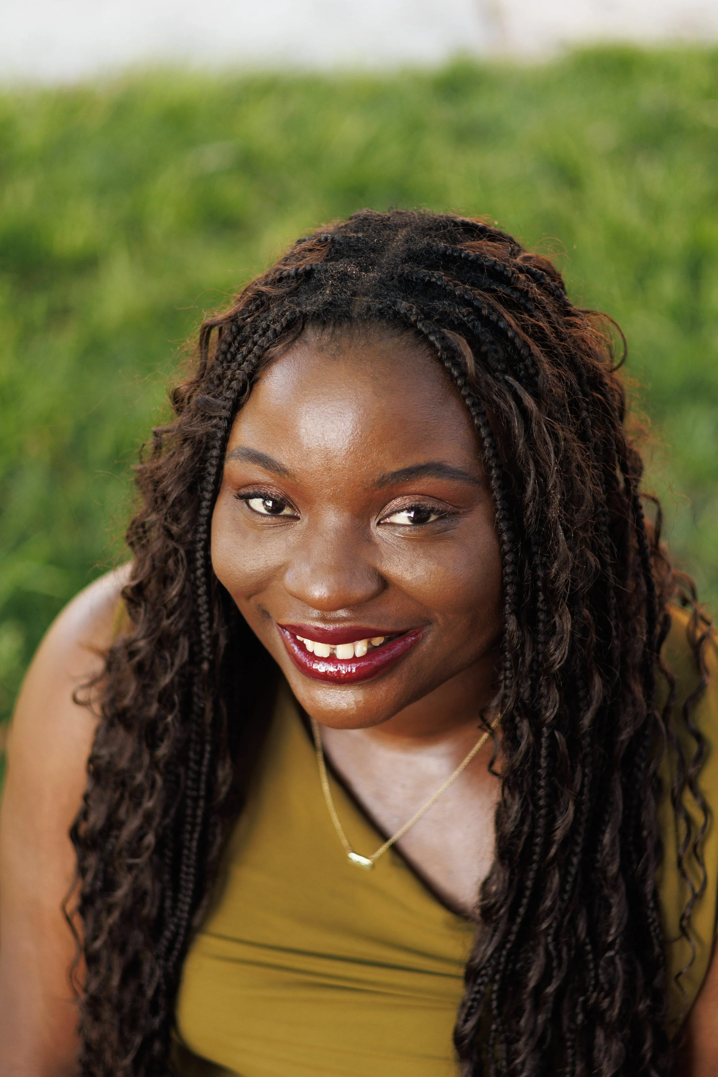 A smiling woman with long braids, wearing a yellow top and a gold necklace, outdoors with a blurred green background.