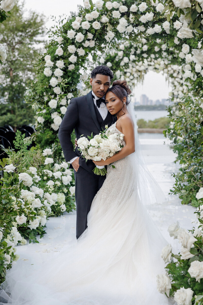 A bride and groom standing closely together under a floral arch at their outdoor wedding. The bride is holding a bouquet of white roses and wearing a white wedding dress with lace and tulle. The groom is dressed in a black tuxedo with a bow tie. The background shows water and a city skyline in the distance.