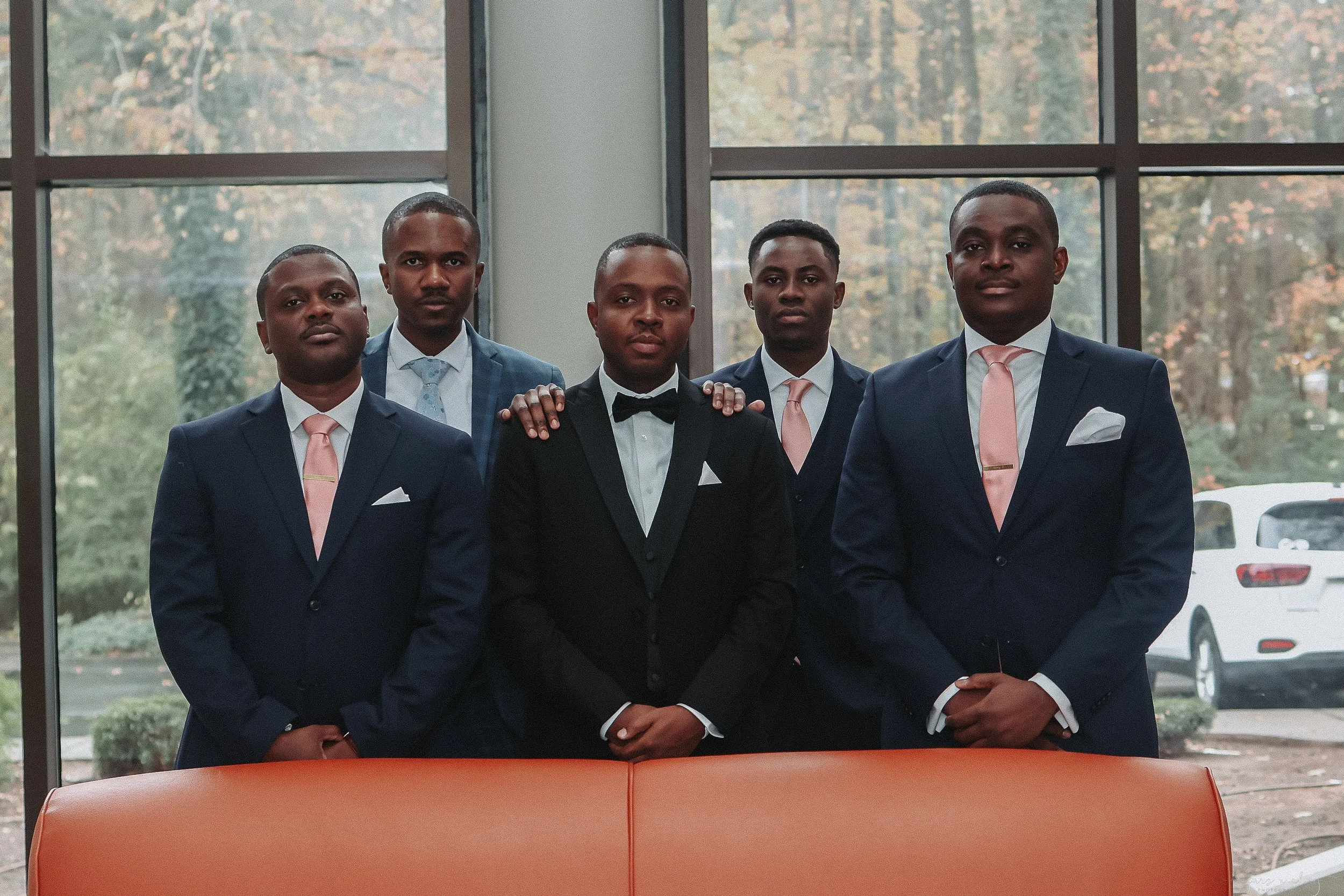 A group of five men dressed in formal suits, standing inside near large windows with an outdoor background of trees and cars.