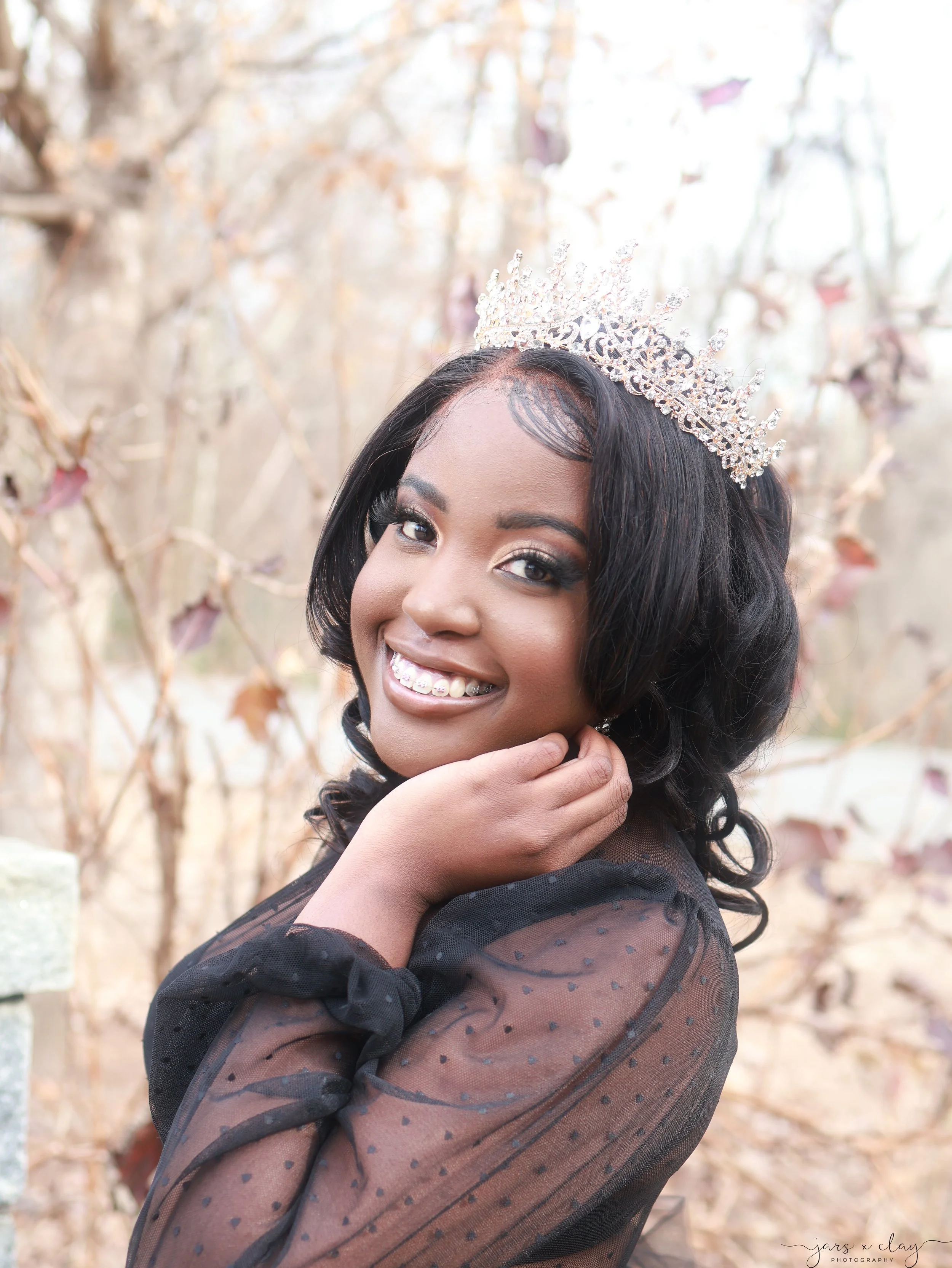 Close-up of a woman with dark hair wearing a tiara and a black, sheer polka dot dress, smiling outdoors with a blurred background of trees and plants.