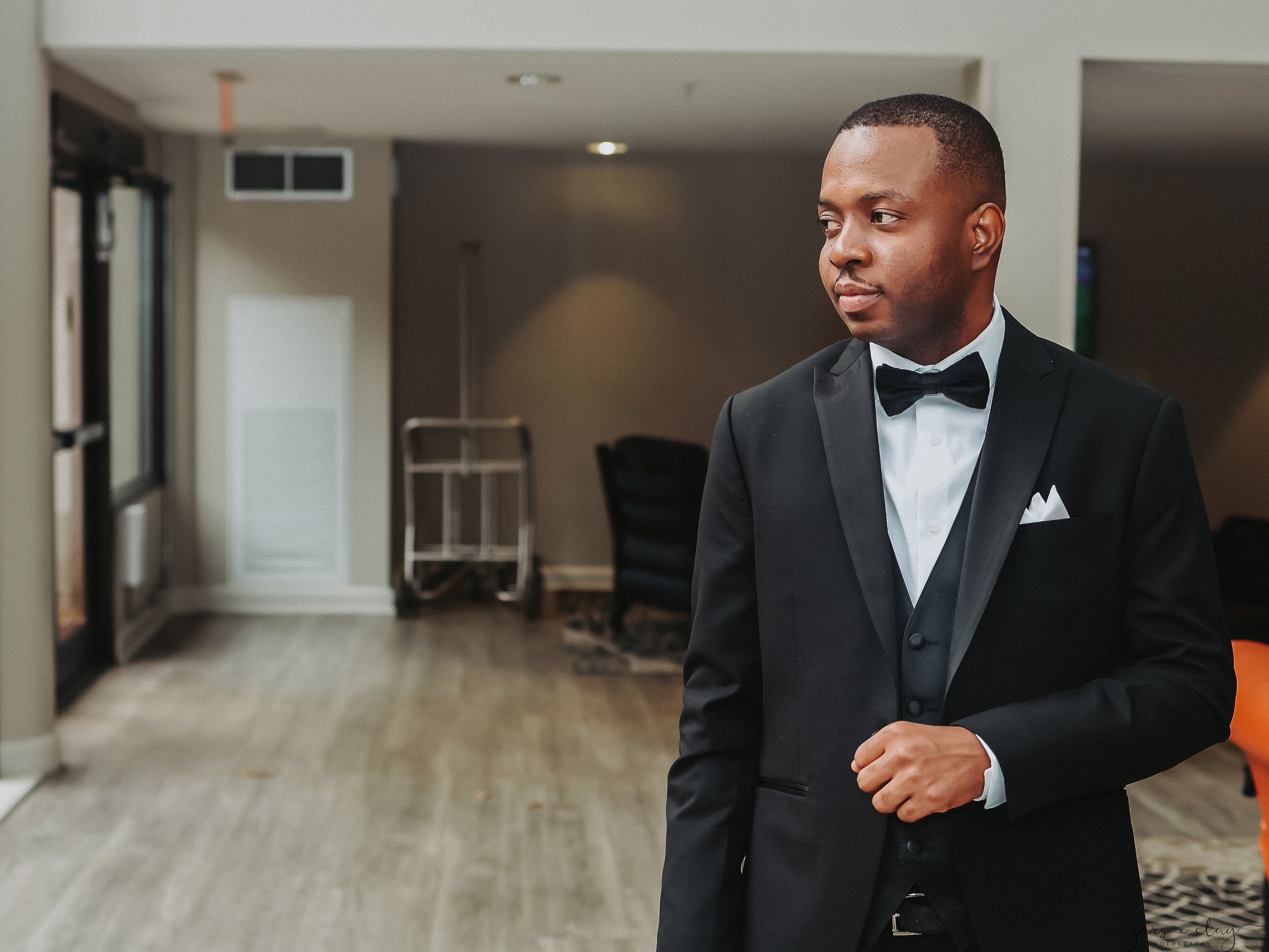 A man dressed in a black tuxedo with a bowtie, standing indoors and looking to his right.