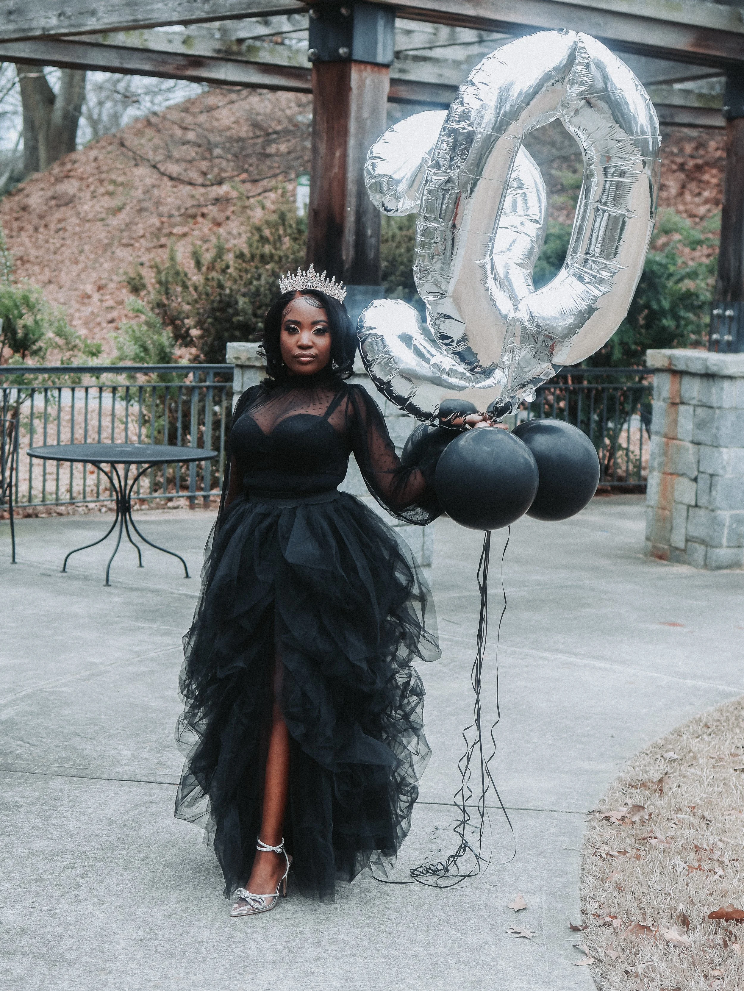 A woman dressed in a black gown with puffy tulle skirt, wearing silver high heels, standing outdoors on a paved surface. She is holding silver and black balloons, with a large silver number 2 balloon visible. She wears a silver tiara and has dark wav