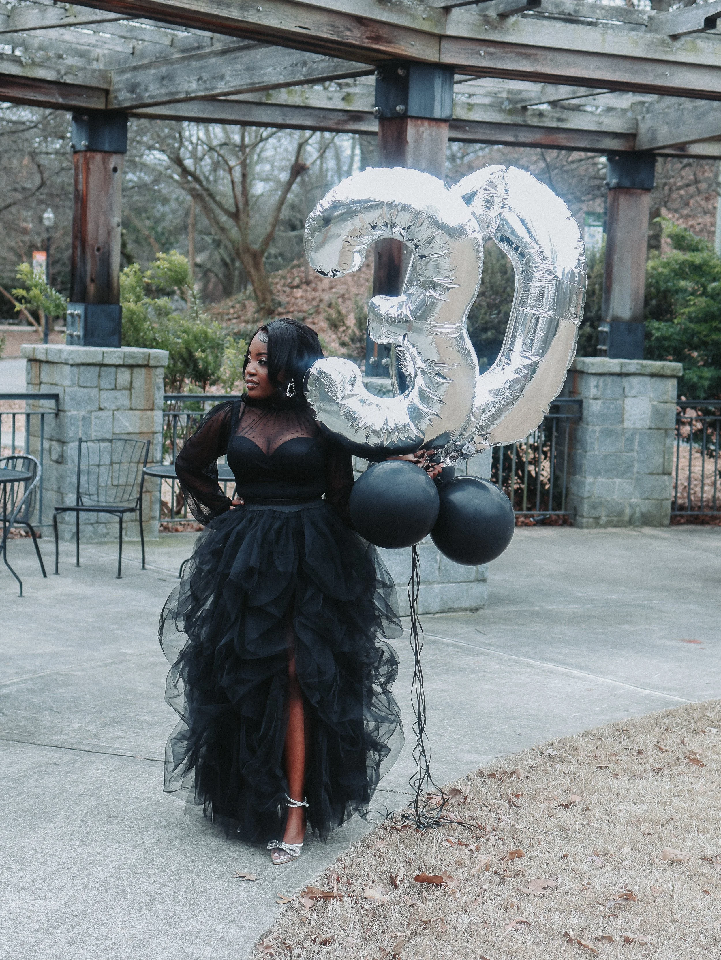 A woman dressed in a black gown holding silver foil balloons shaped as the number three and the number zero, standing outdoors beneath a wooden structure with stone pillars, with trees and chairs in the background.