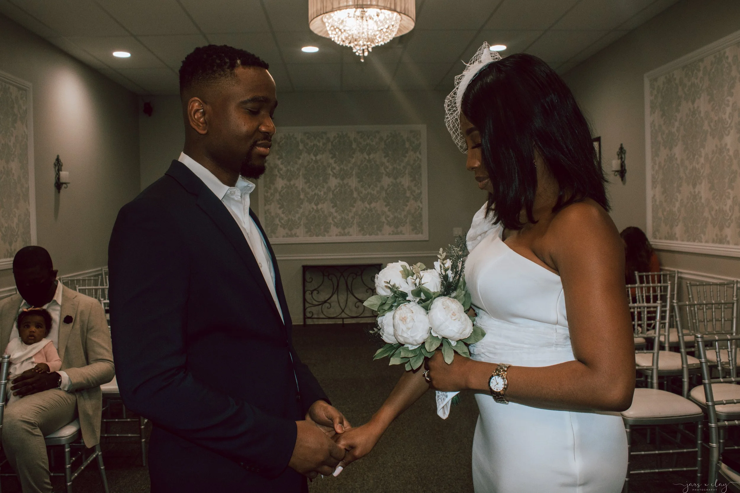 An African American couple holding hands during a wedding ceremony in a decorated indoor venue, with seated guests observing.
