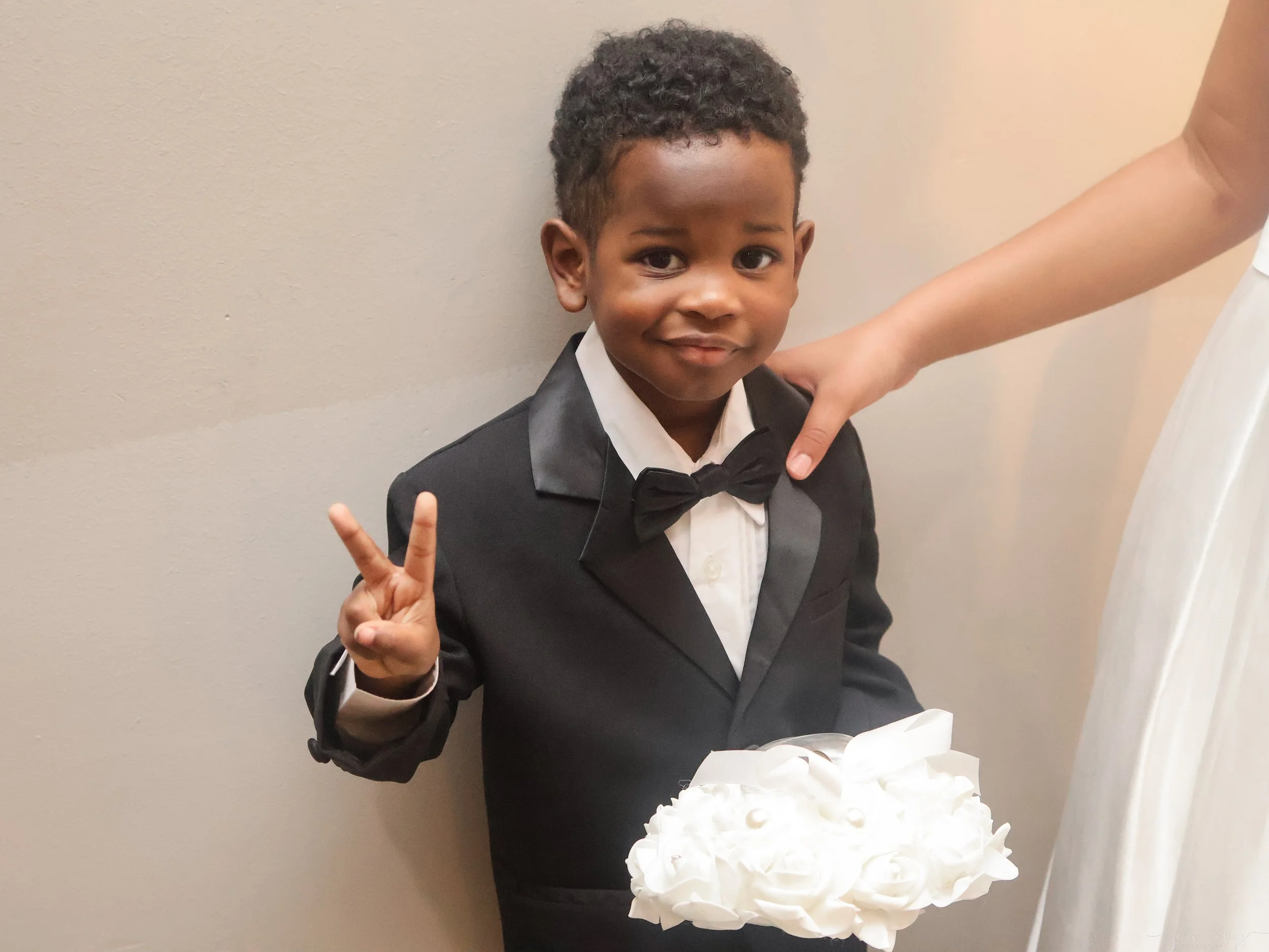 Young boy in a tuxedo making a peace sign with his hand, standing behind a white cake decorated with whipped cream.