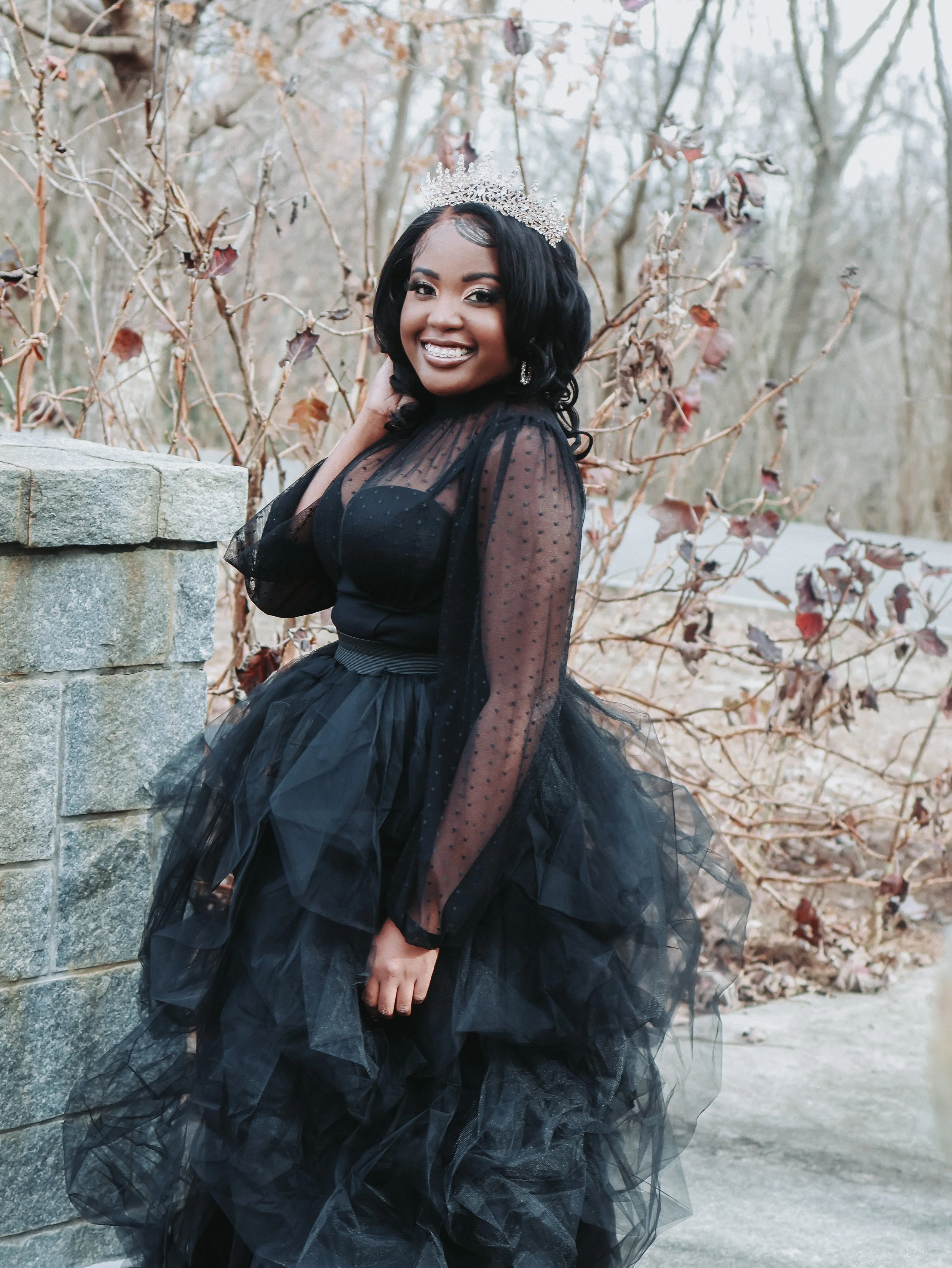 A woman with dark hair and dark skin wearing a black dress with sheer polka-dot sleeves and a voluminous black tulle skirt, smiling and showing her teeth, wearing a silver tiara, standing outdoors near a stone wall and leafless bushes.