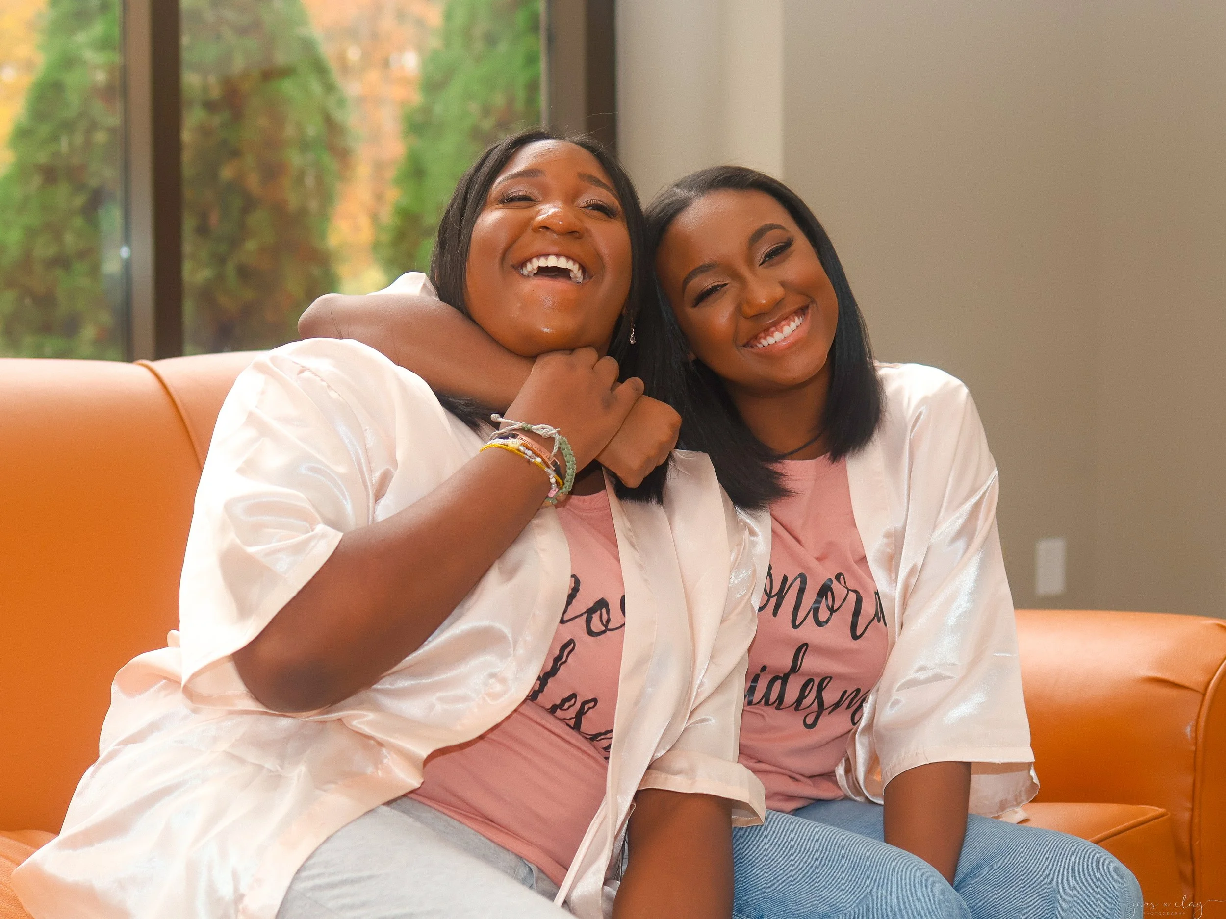 Two women sitting on a beige couch, smiling and hugging each other warmly, with a window showing fall foliage in the background.