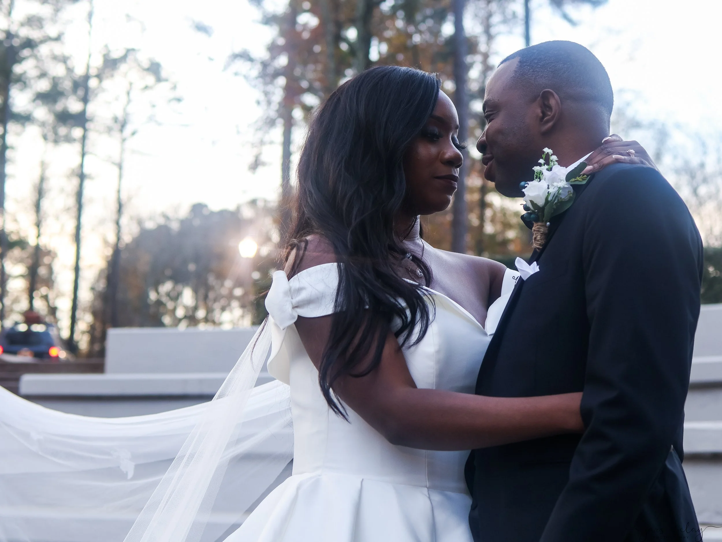 A couple in wedding attire, with the woman wearing a white dress and the man in a dark tuxedo, embrace each other outdoors in front of trees during sunset.