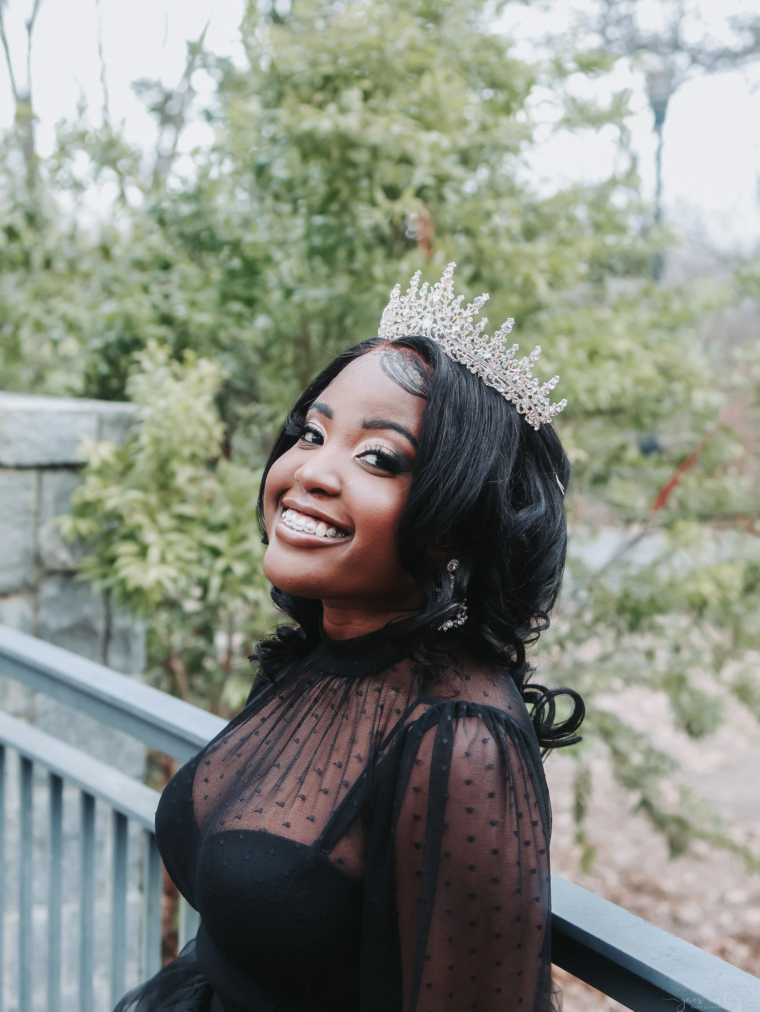 A Black woman with black, wavy hair wearing a silver tiara and black dress with sheer polka dot sleeves, smiling outdoors near a gray railing and trees.