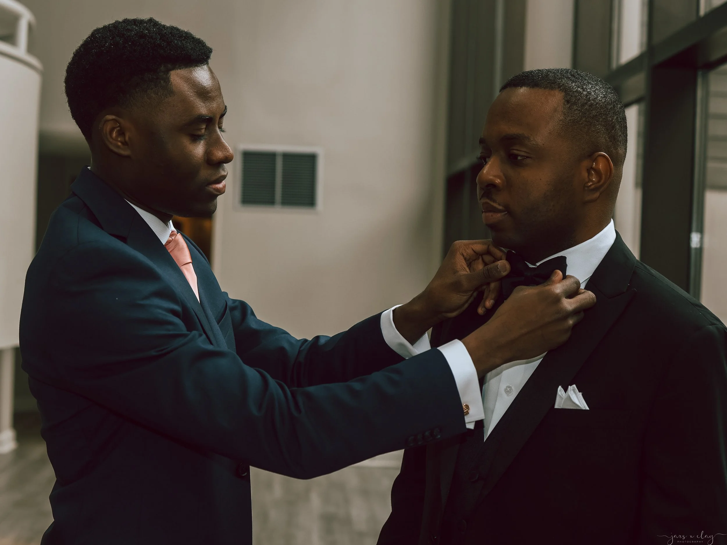Two men in tuxedos are in a narrow hallway, with one helping the other adjust his bow tie. They are dressed formally, suggesting a special event or occasion.
