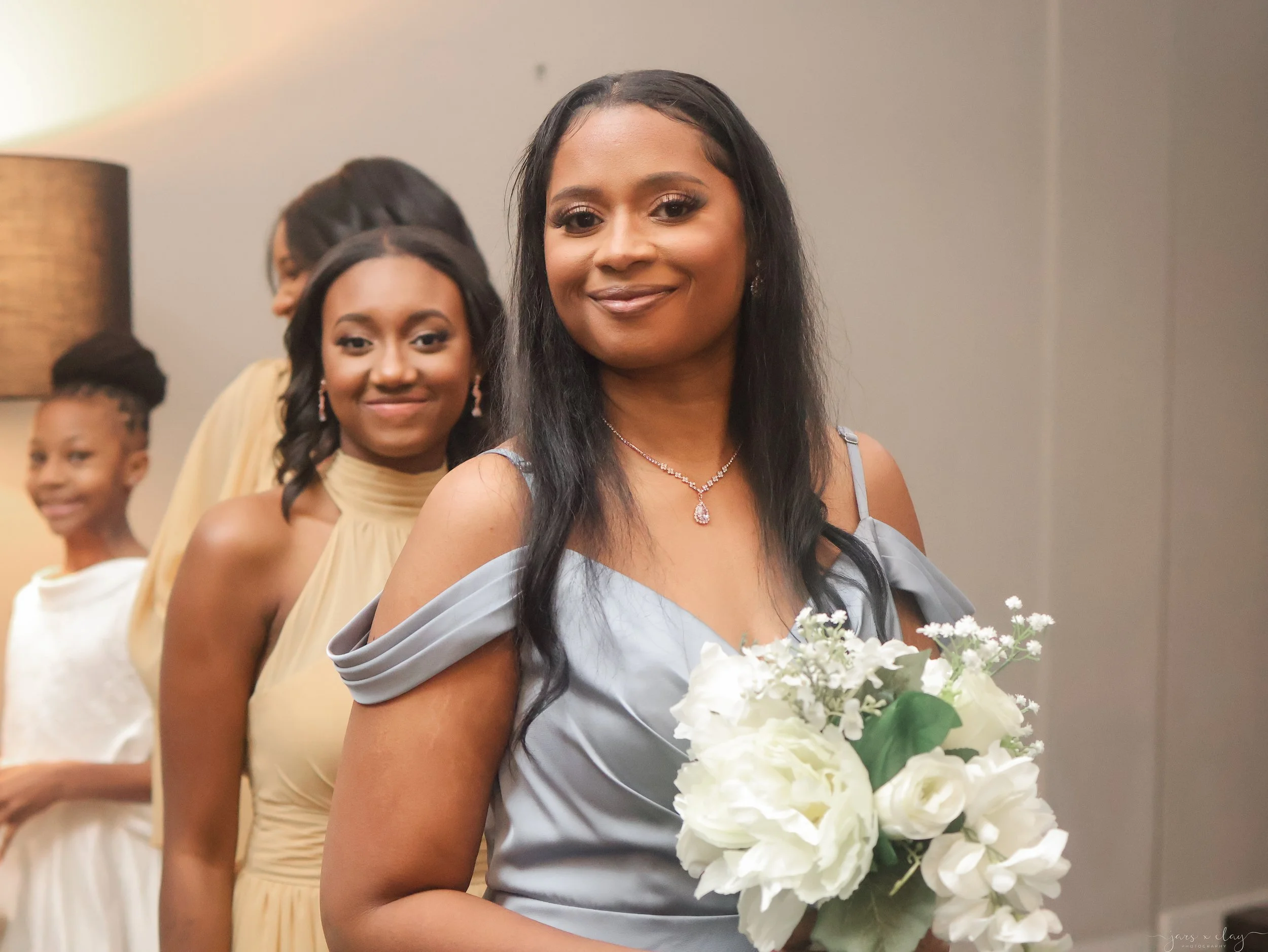 Women dressed in formal attire standing in line, with the woman in the foreground holding a bouquet of white flowers and wearing jewelry, at what appears to be a wedding or celebration event.