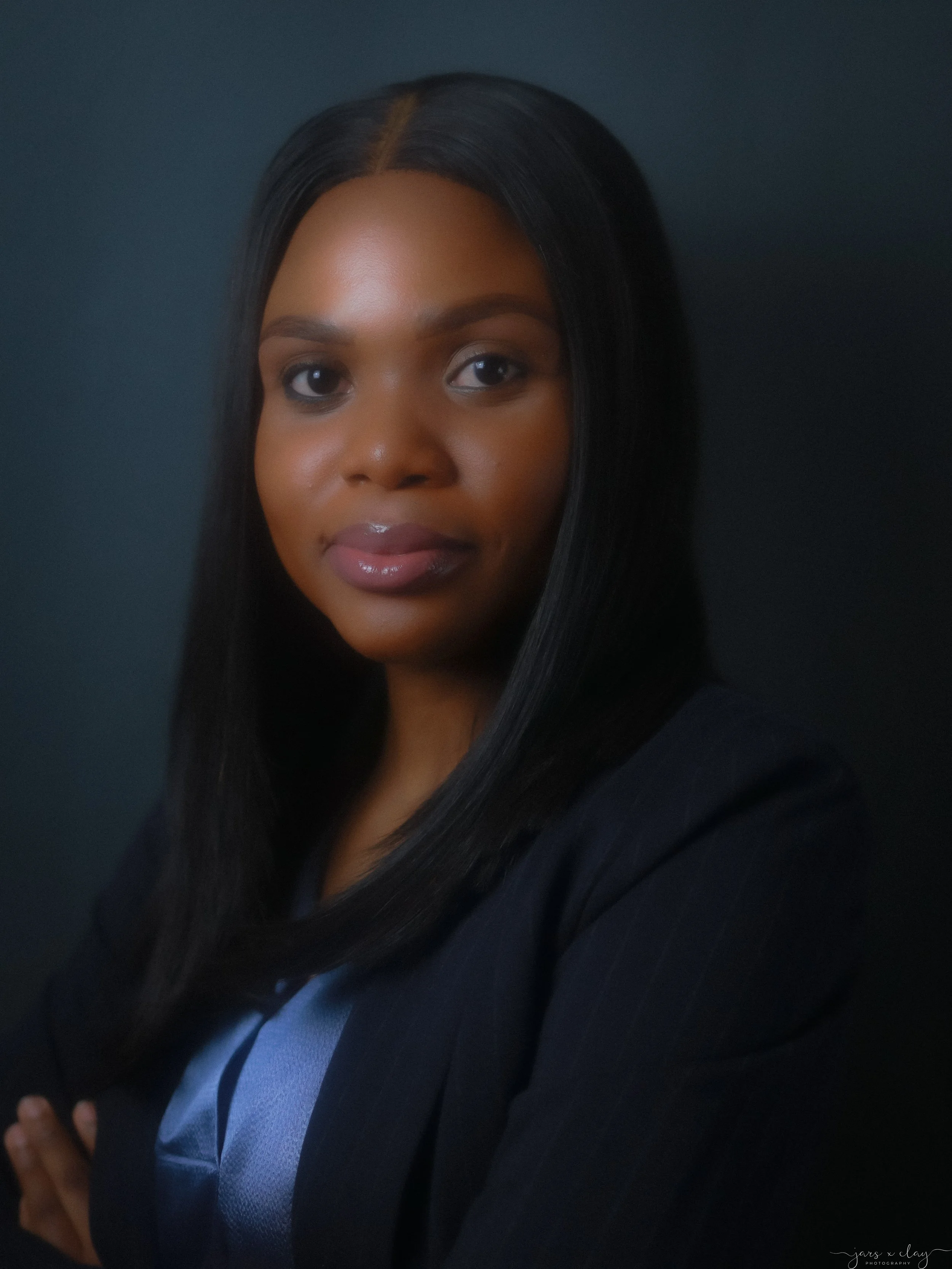 Close-up portrait of a woman with dark hair, wearing a dark blazer and a light-colored top, posing against a dark background.