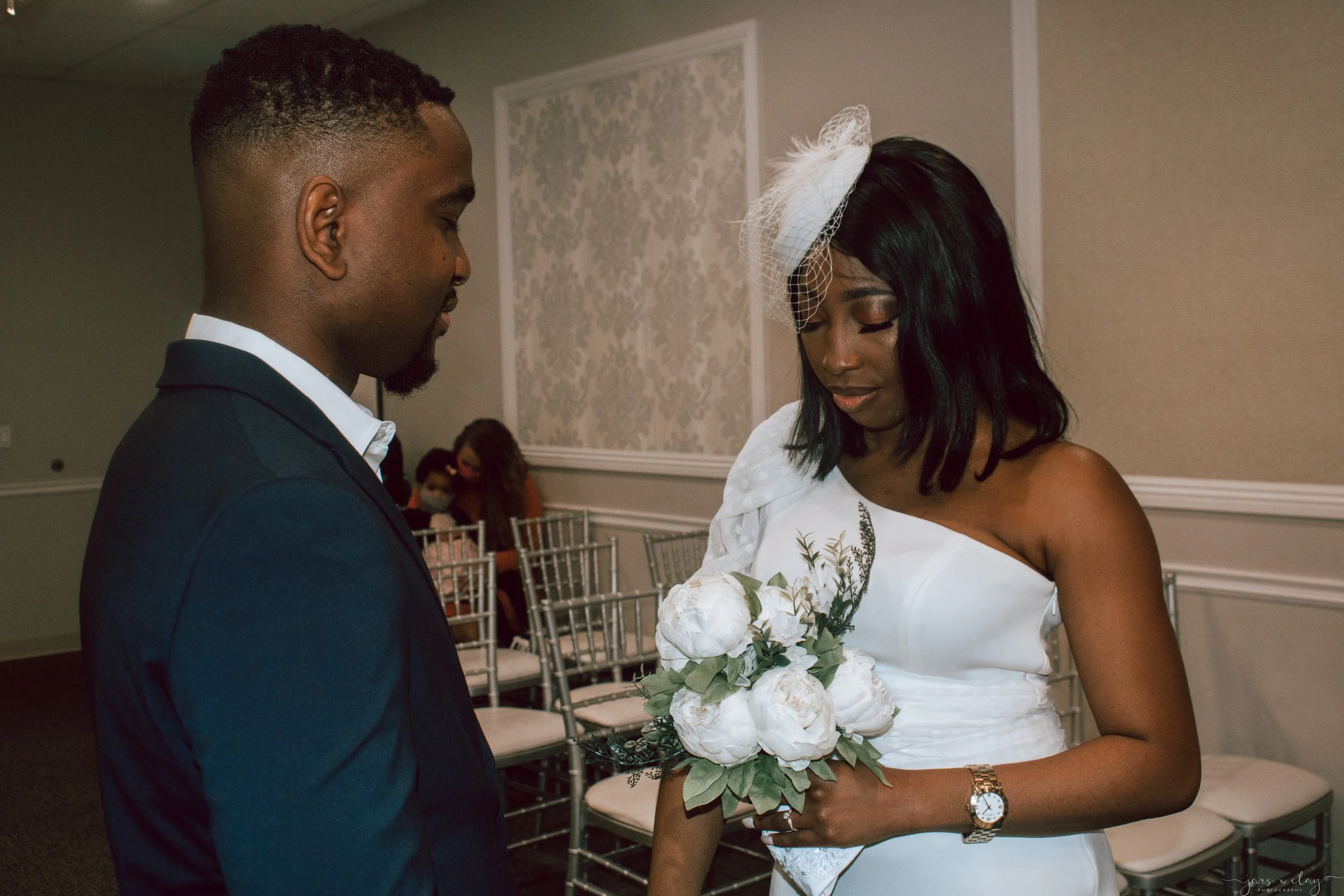 A bride and groom are sharing a moment during their wedding ceremony. The bride is holding a bouquet of white flowers and wearing a white dress with a fascinator. The groom is dressed in a dark suit.