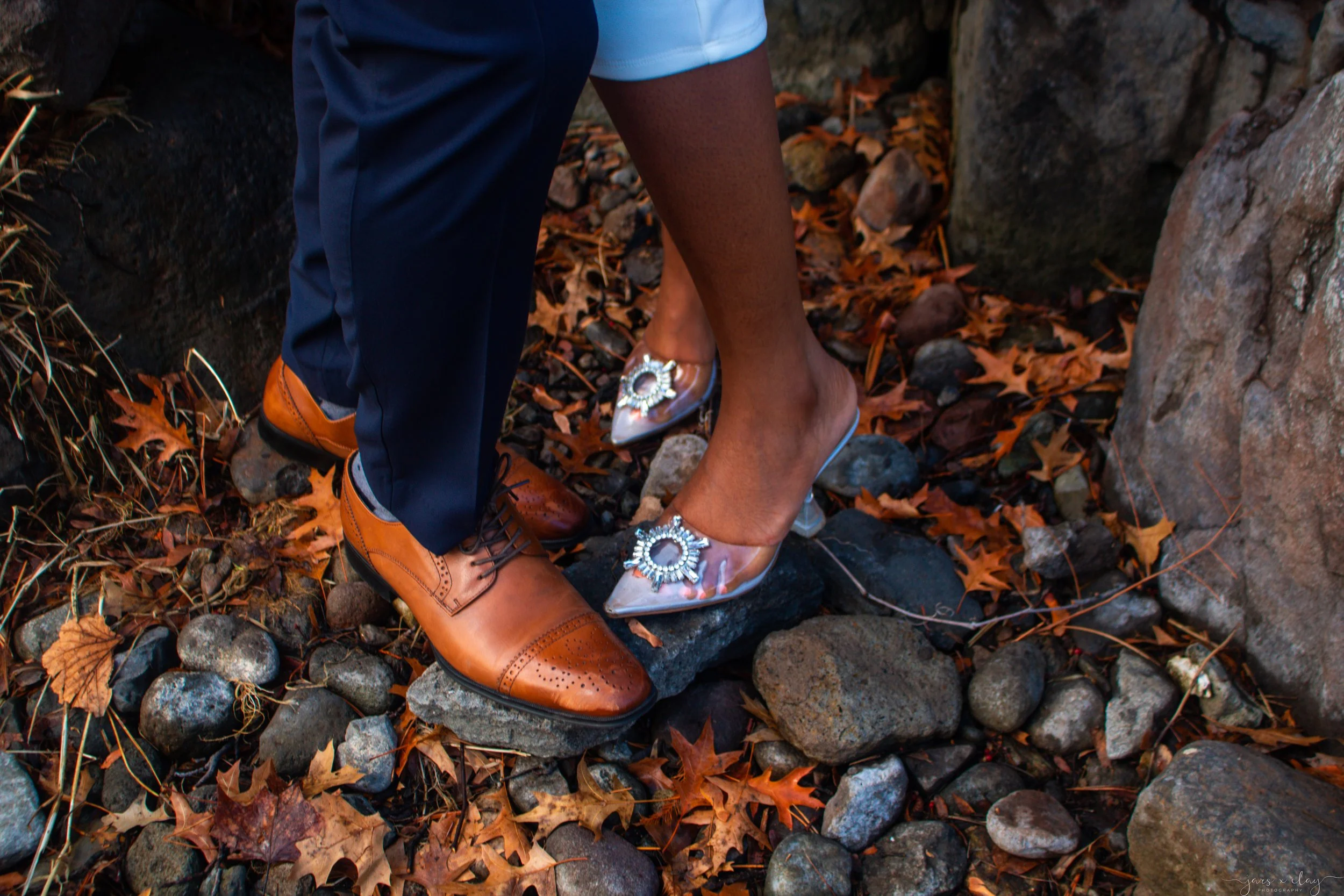 A person wearing brown dress shoes, dark blue pants, and blue shorts is standing on a rocky surface surrounded by fallen autumn leaves. They are also wearing transparent high-heeled shoes with decorative embellishments.