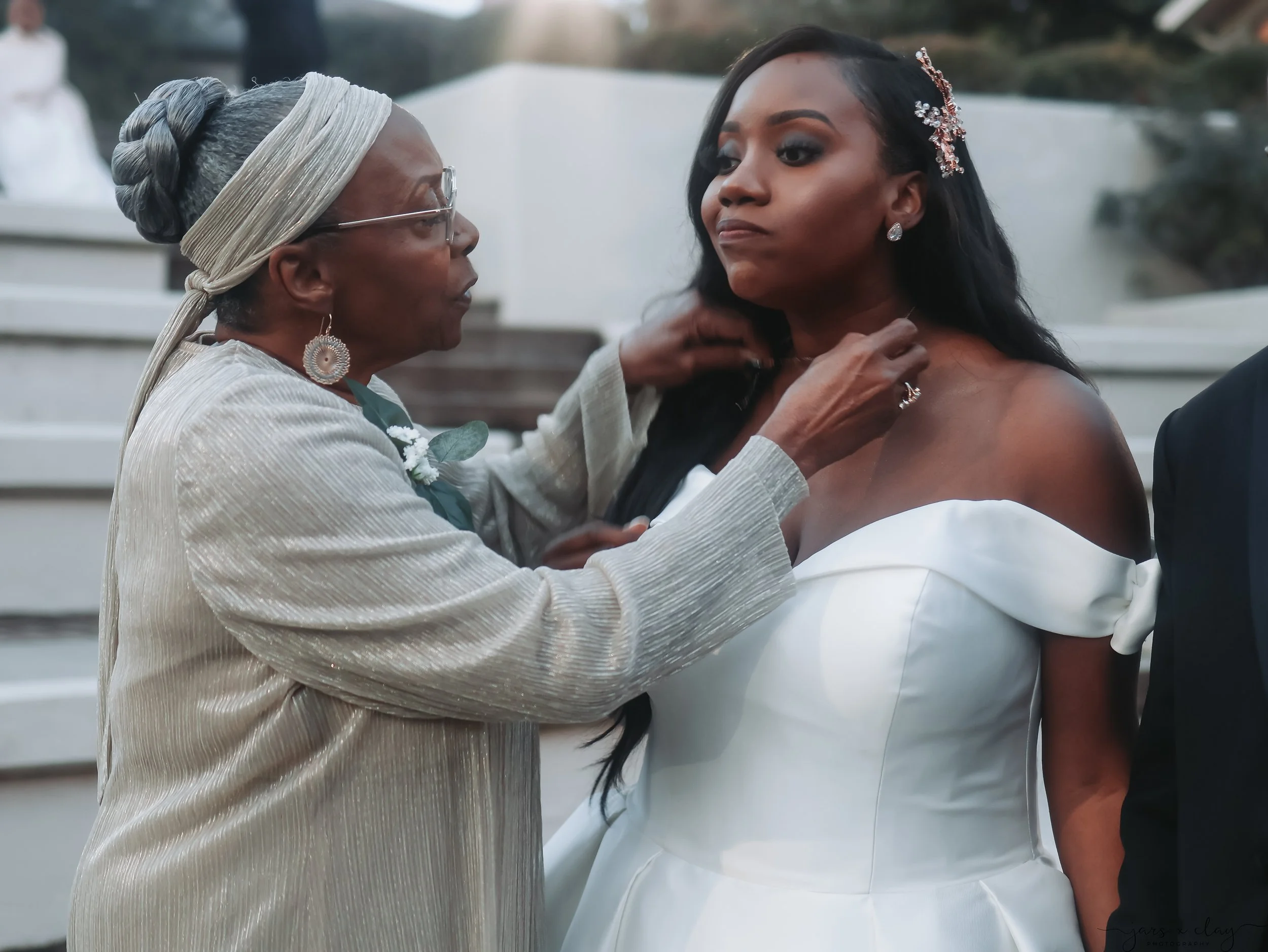 An elderly woman with glasses and gray hair styled in an updo, wearing a cream-colored, textured dress and large earrings, is adjusting the strap of a young woman's off-the-shoulder white dress, likely at a wedding or special event outdoors with a wh