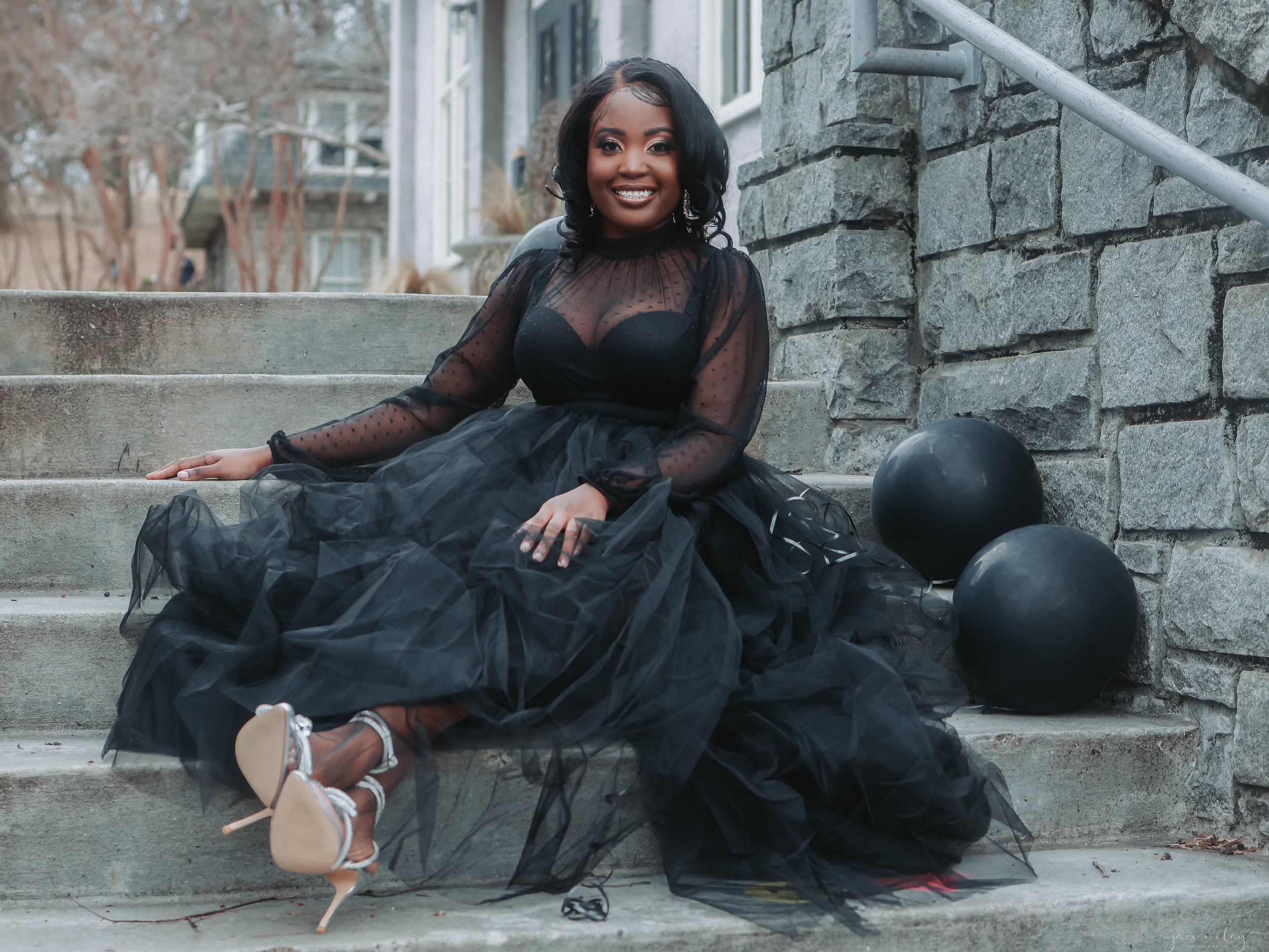 A woman sitting on concrete stairs outdoors, smiling, wearing a black sheer top and black tulle skirt, with balloons around her. Background includes stone wall, house, and trees.