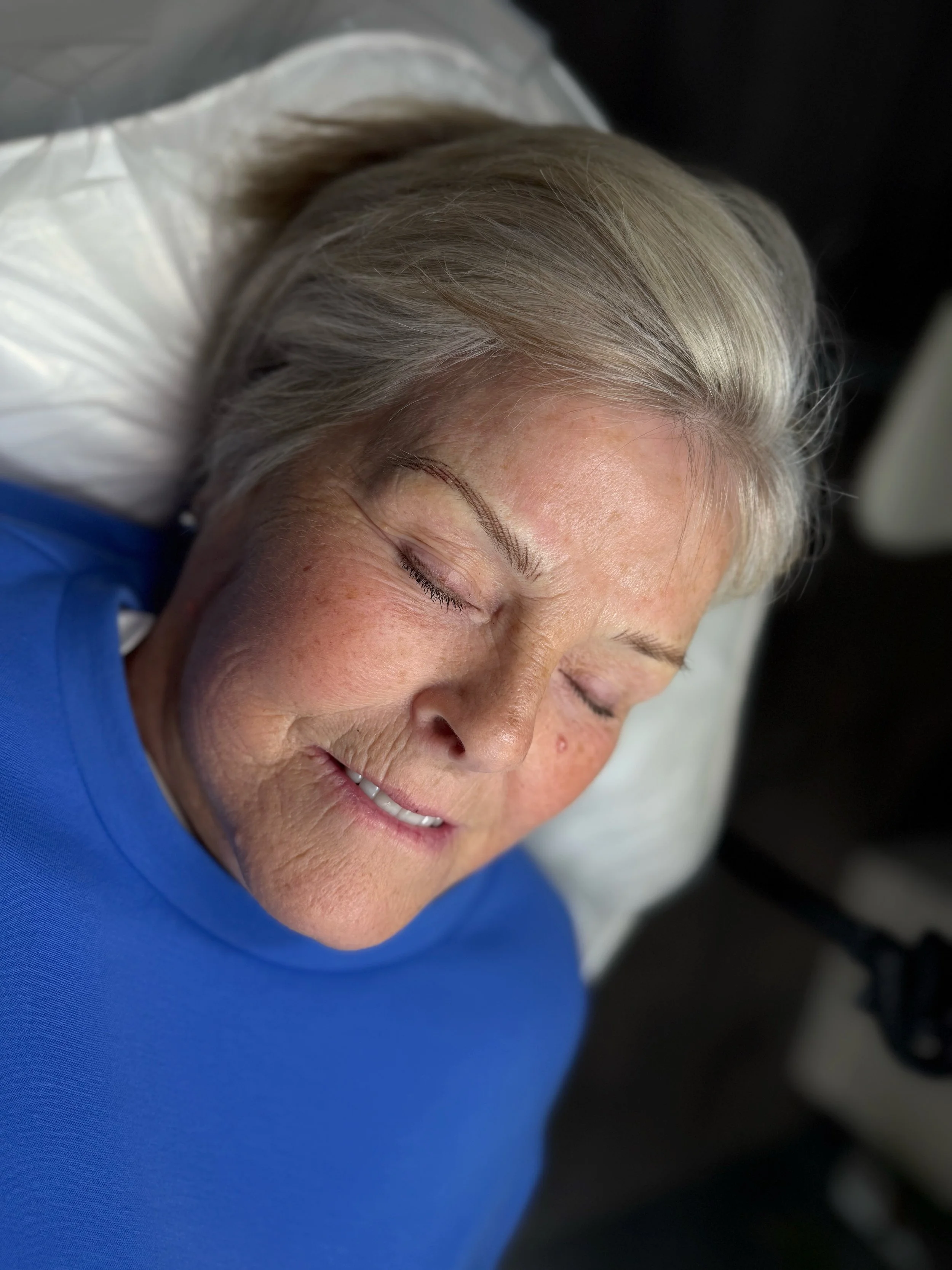 A close-up of an elderly woman with gray hair sleeping peacefully with her head resting on a pillow, wearing a blue shirt.