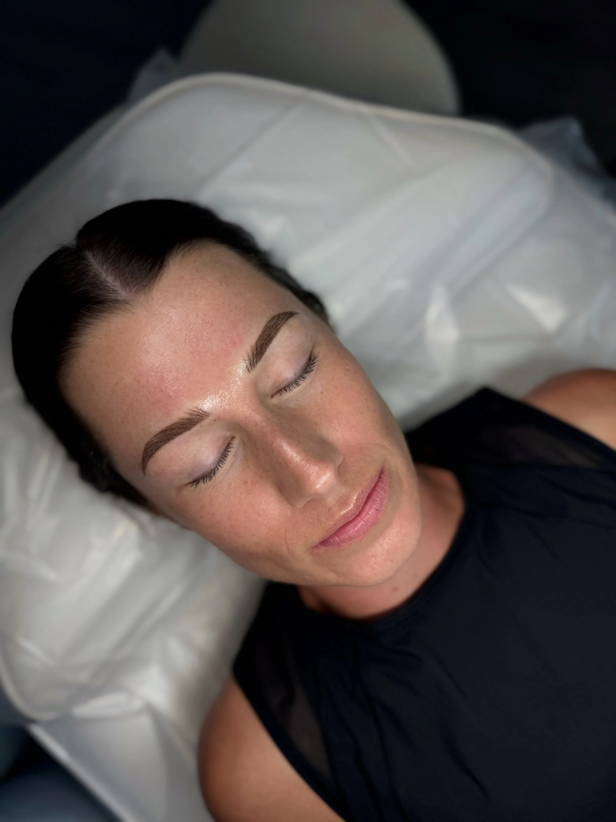 A woman with dark hair and neatly shaped eyebrows lying on a pillow with her eyes closed, appearing relaxed.