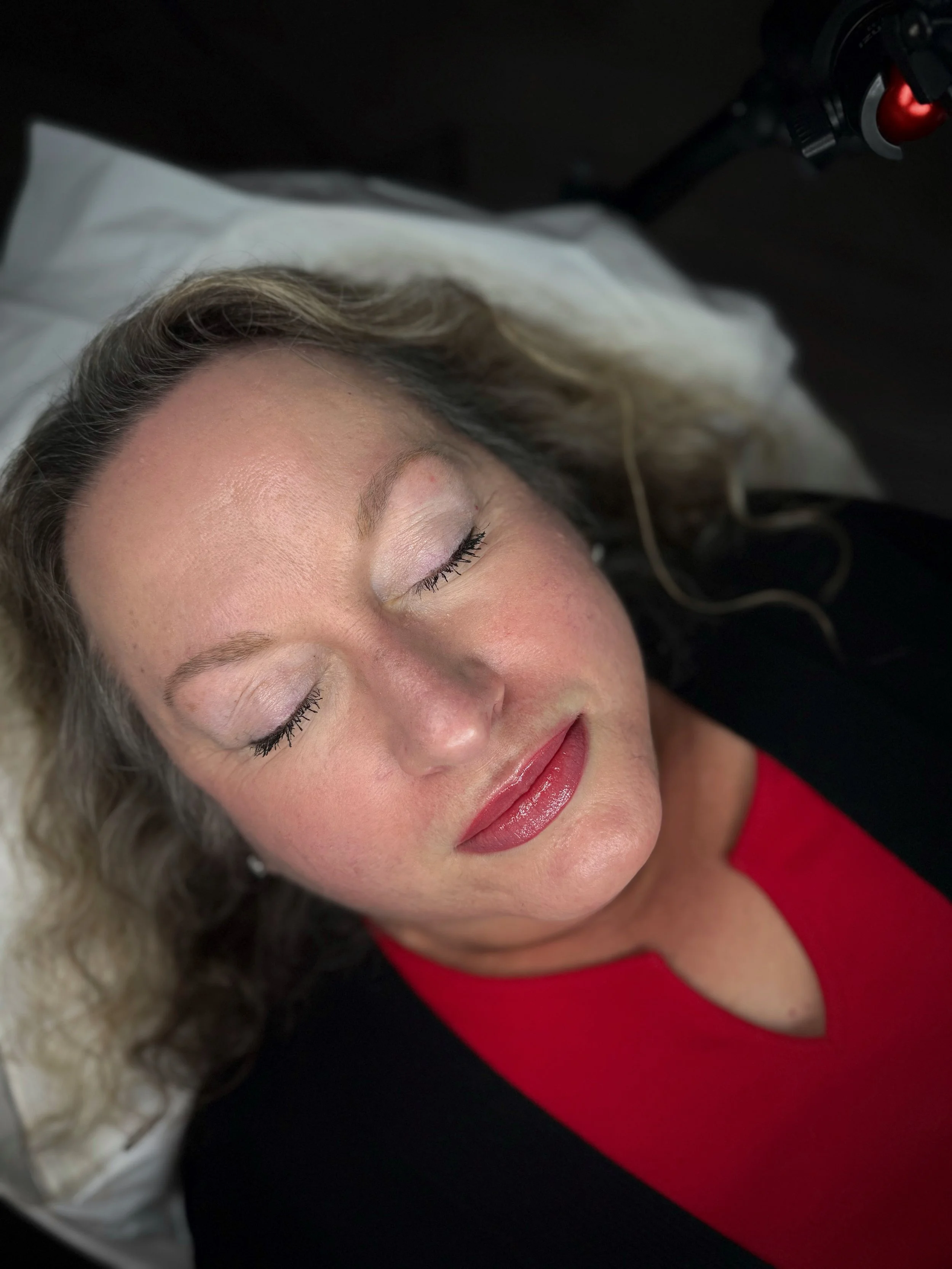 A woman with blonde hair and makeup, lying on her back with eyes closed, wearing a red top and a black outer garment.