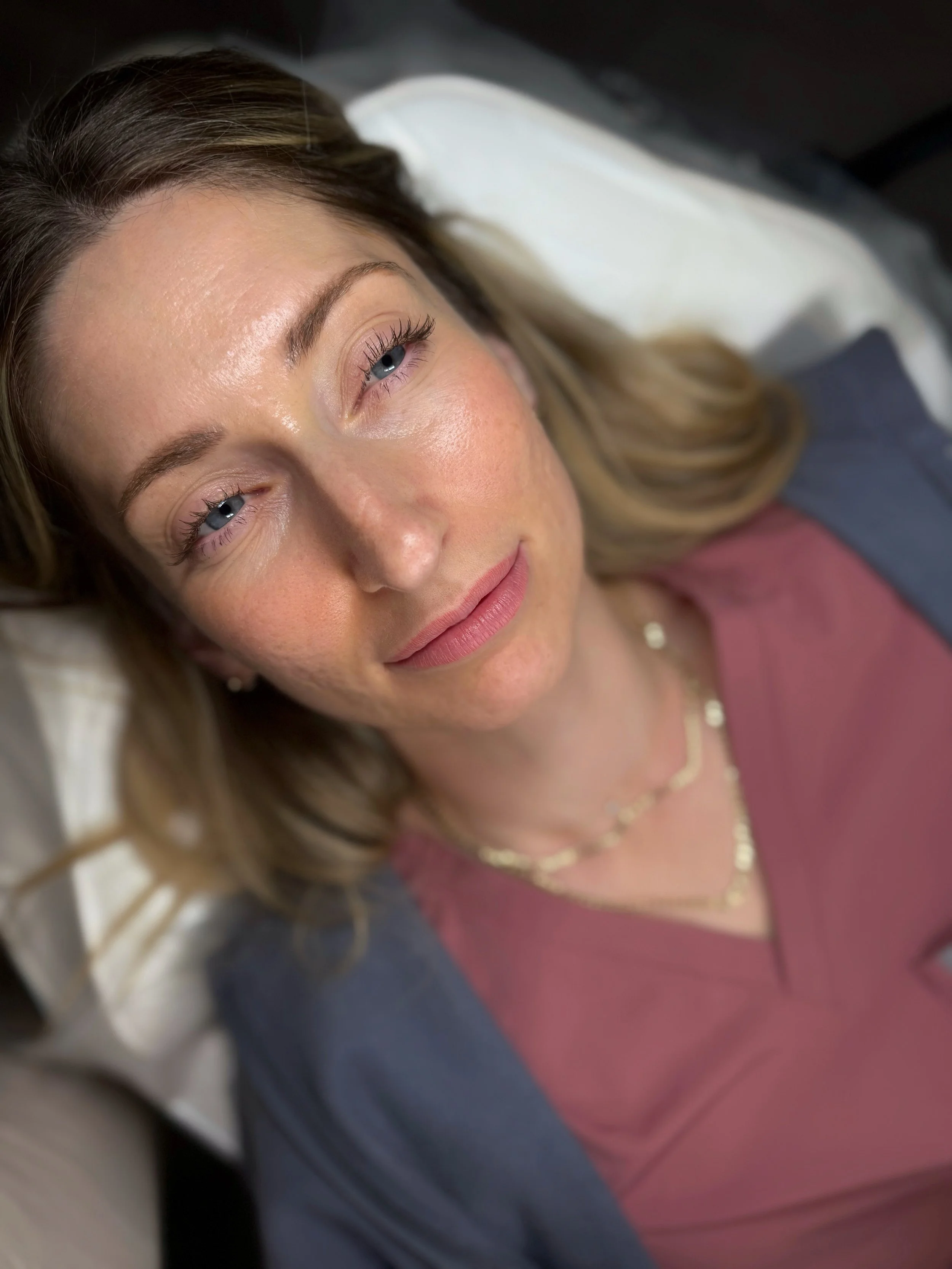 Close-up of a woman with blue eyes and light makeup, lying on a pillow with a relaxed expression, wearing a pink top and layered necklaces.