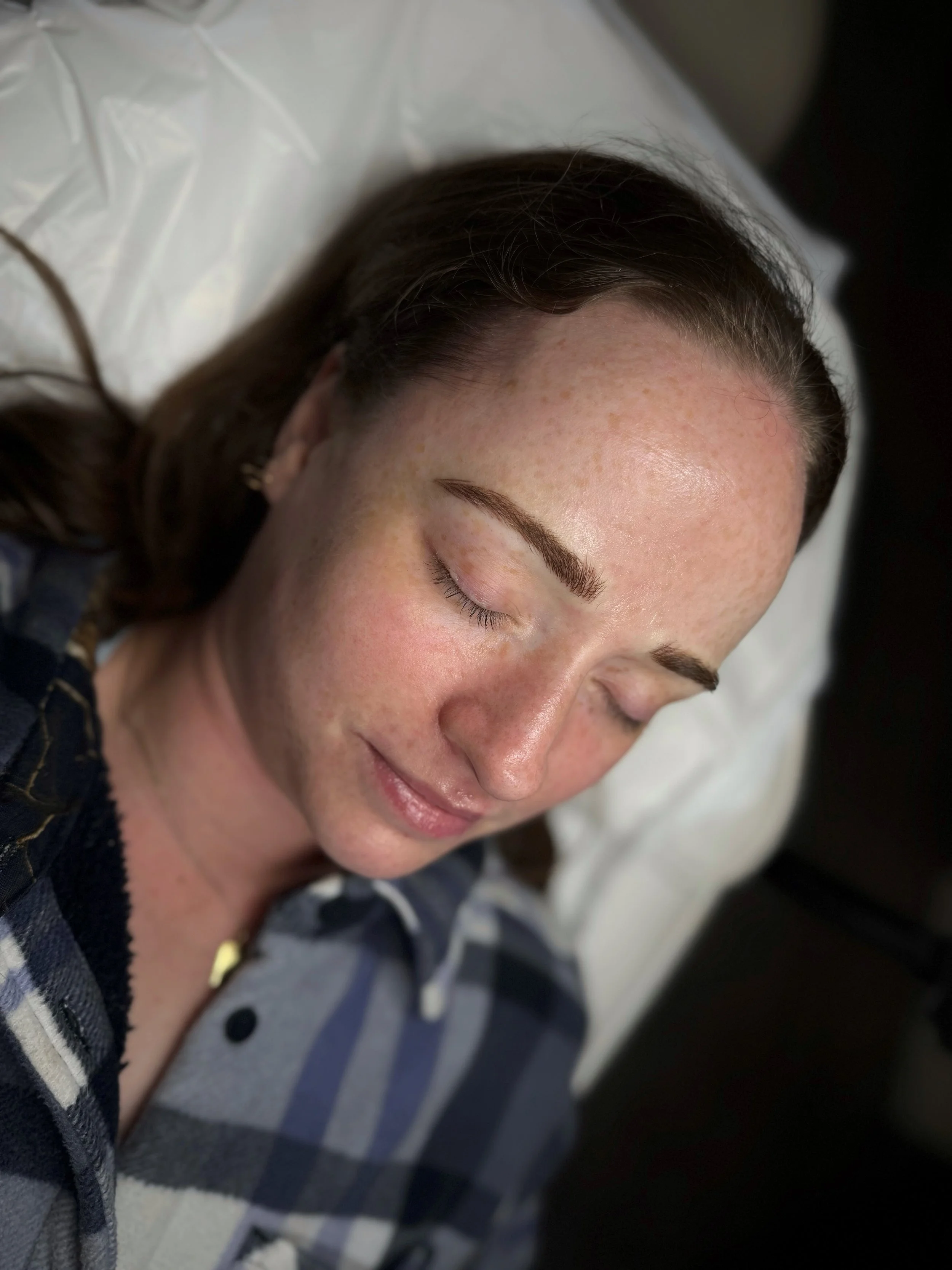 A woman with closed eyes, lying on a pillow with a relaxed smile, has freckles on her face, and is wearing a plaid shirt.