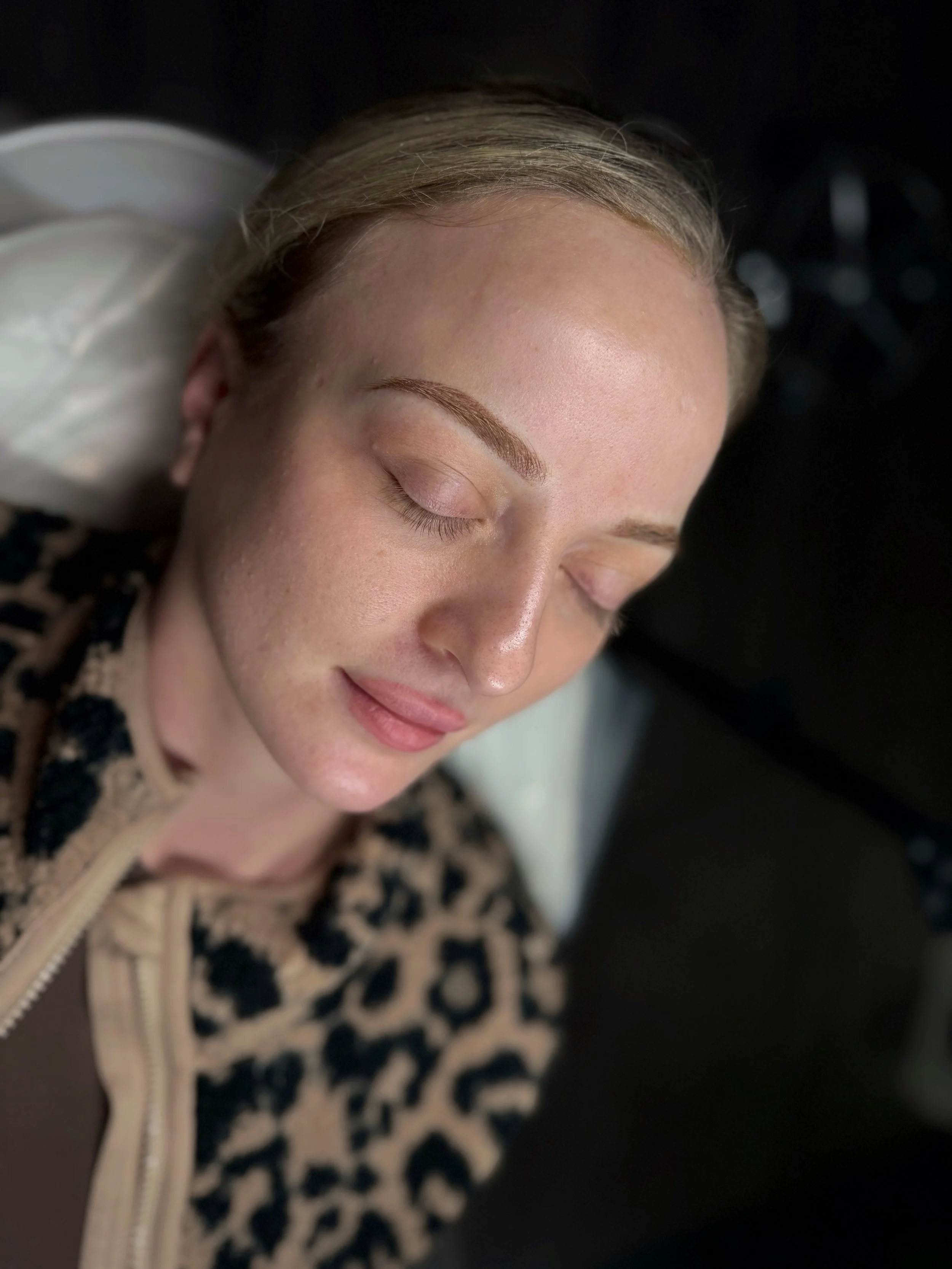 Young woman with closed eyes and relaxed expression lying down, wearing a leopard print top, freshly tattooed eyebrows