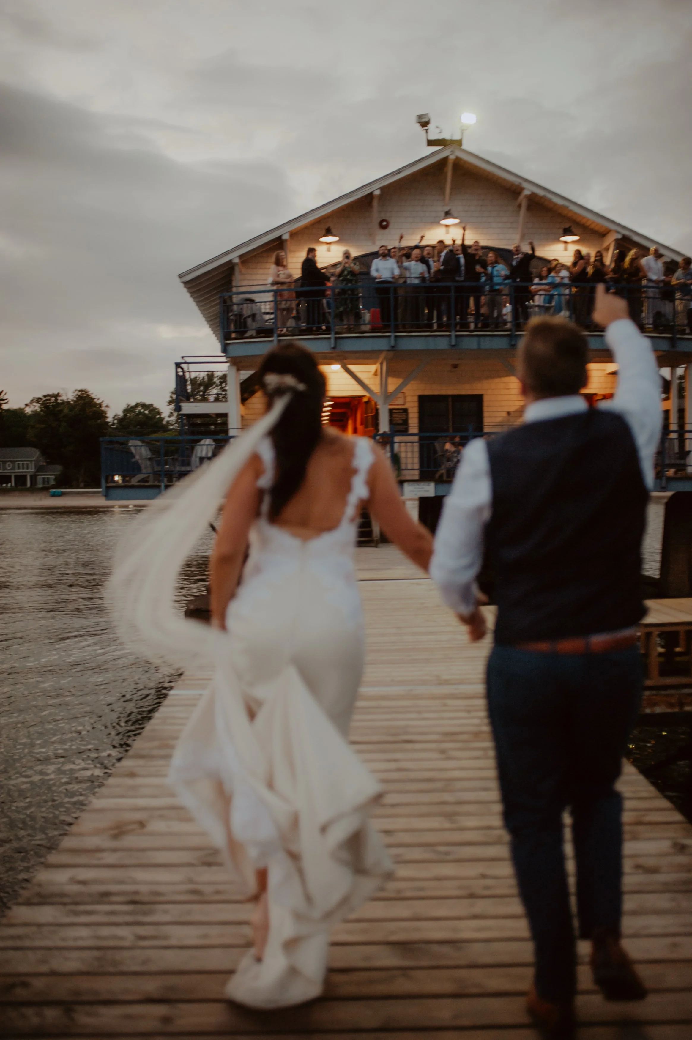 Bride and groom walking on a wooden dock towards a two-story building with guests on the upper balcony, near a body of water, at dusk at the buffalo canoe club in crystal beach, Ontario.