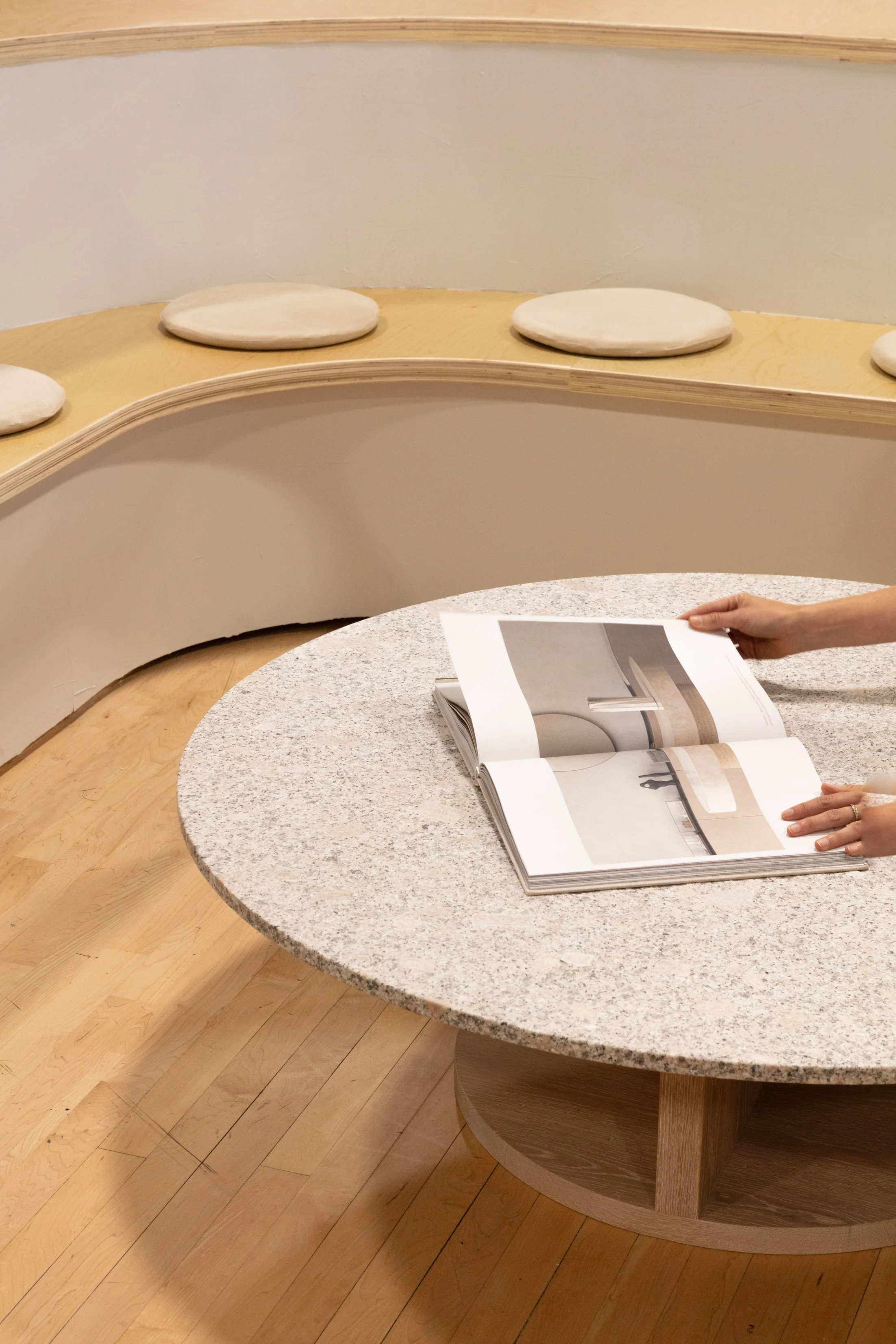 Hands browsing an interior design book on a round granite table in a modern curved seating area