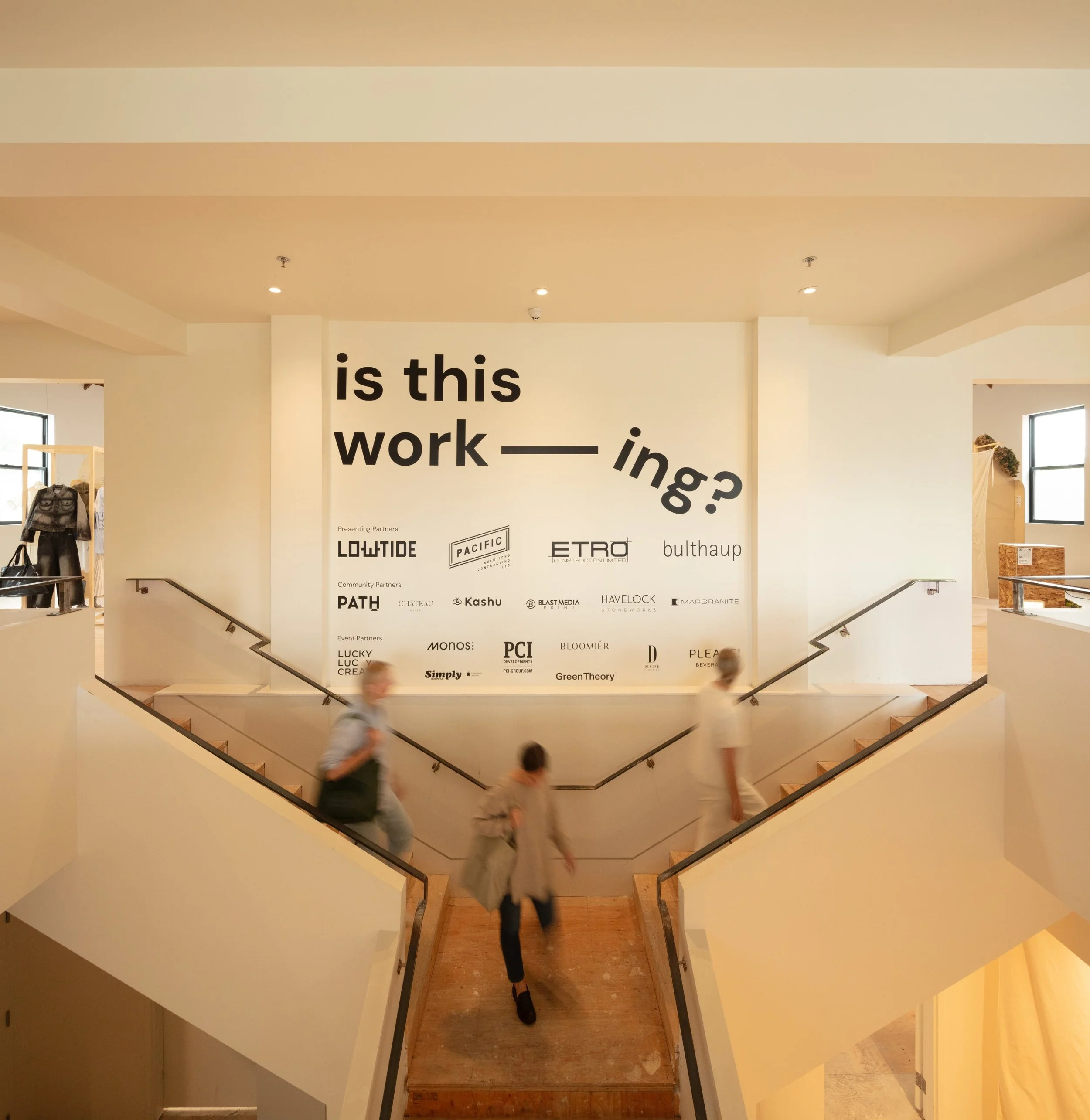 Visitors walking down a staircase in front of a large wall installation reading ‘is this work—ing?’ with sponsor logos